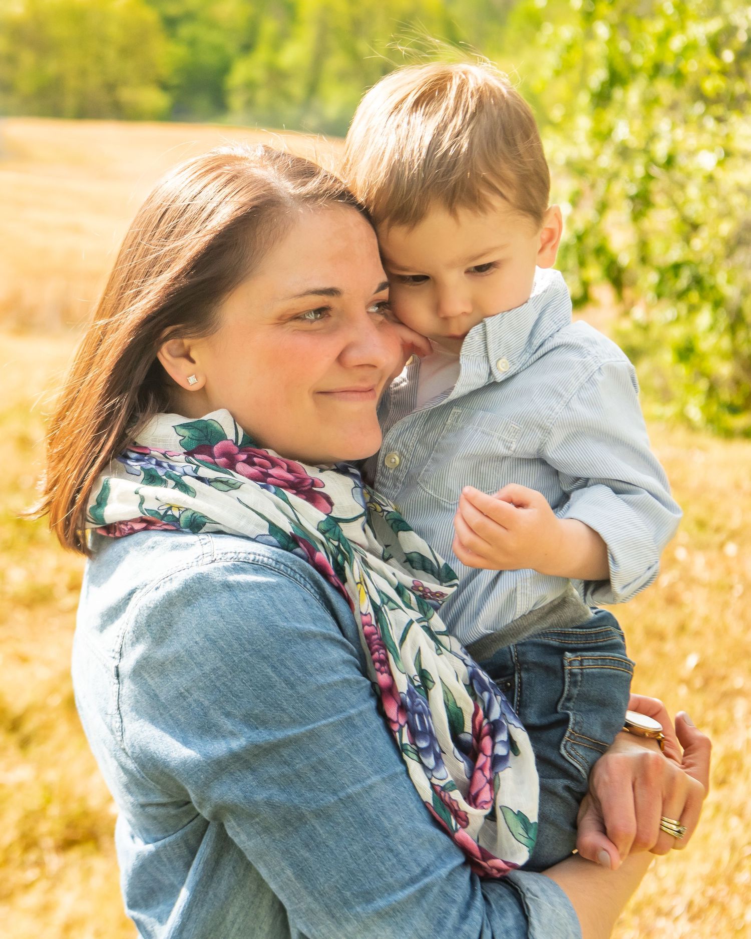 A parent and child share a tender embrace outdoors in golden sunlight while wearing casual denim and a floral scarf.