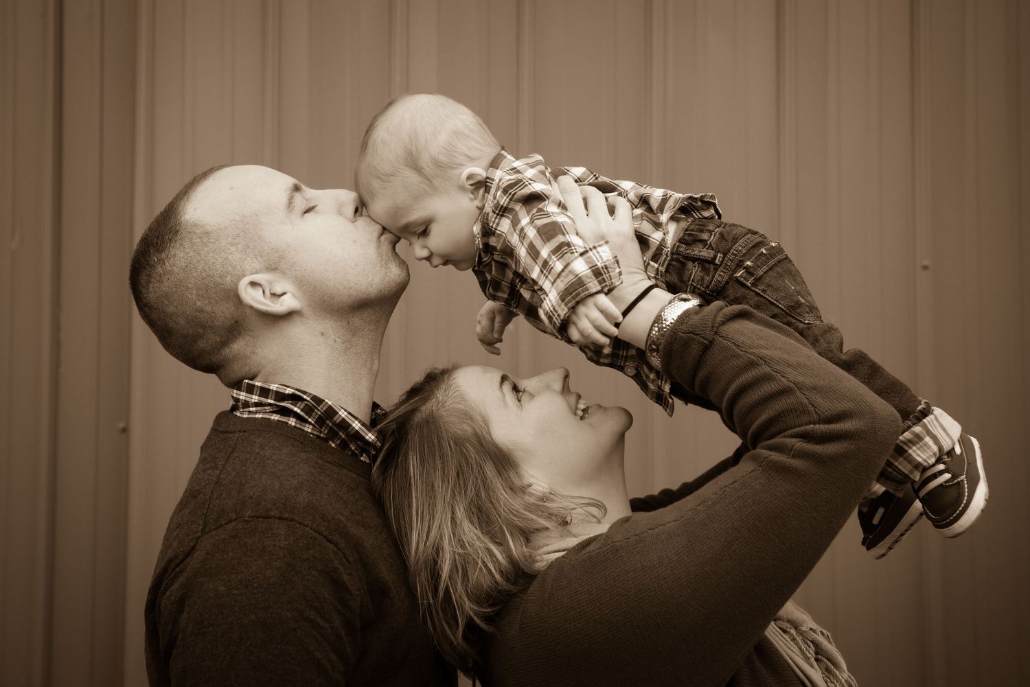 A heartwarming family moment in sepia tones shows parents lifting and kissing their small child.