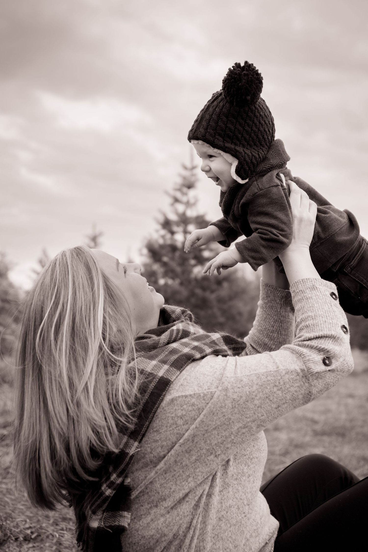 Black and white photo showing a tender moment outdoors as a small child in a winter hat is lifted up in play by his mother.