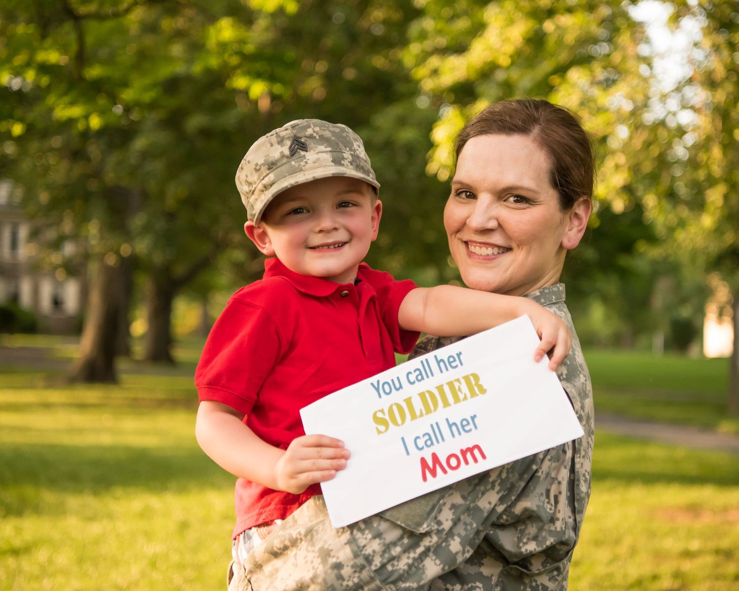 A person in military uniform and a child in a red shirt pose outdoors with a sign in golden sunlight.