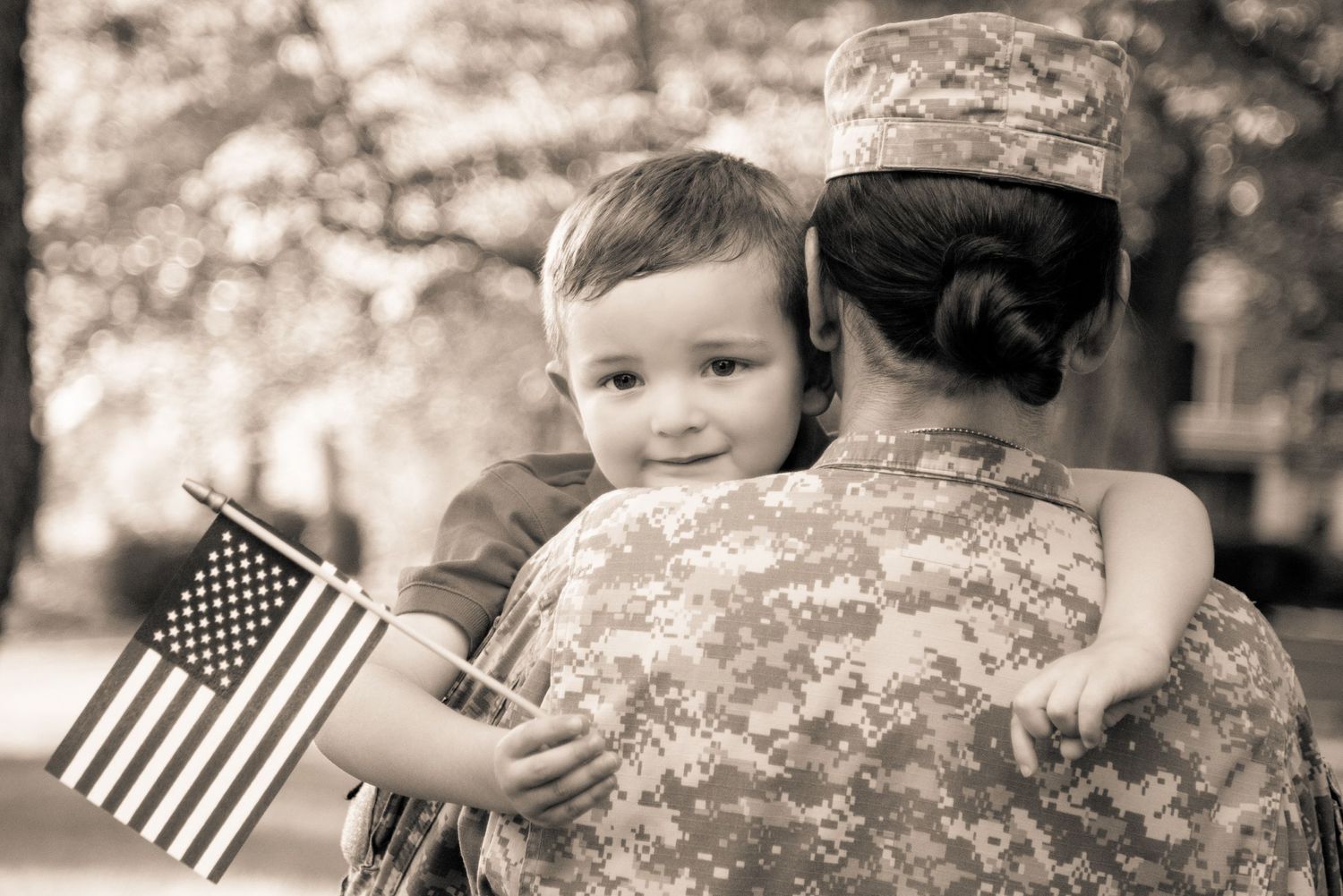 A small child holds an American flag while being held by someone wearing military camouflage in a sepia-toned photograph.