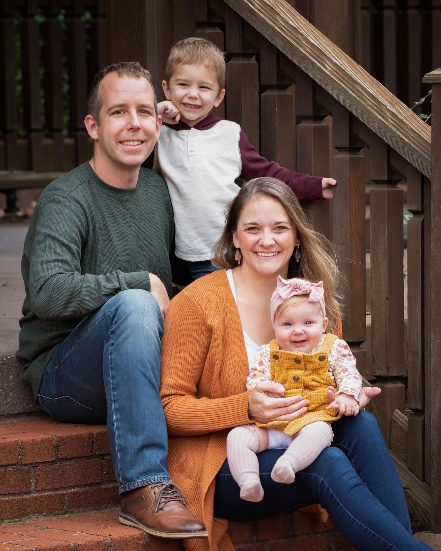 A family sits together on outdoor brick steps with wooden railings, wearing casual autumn clothing in orange and green tones.