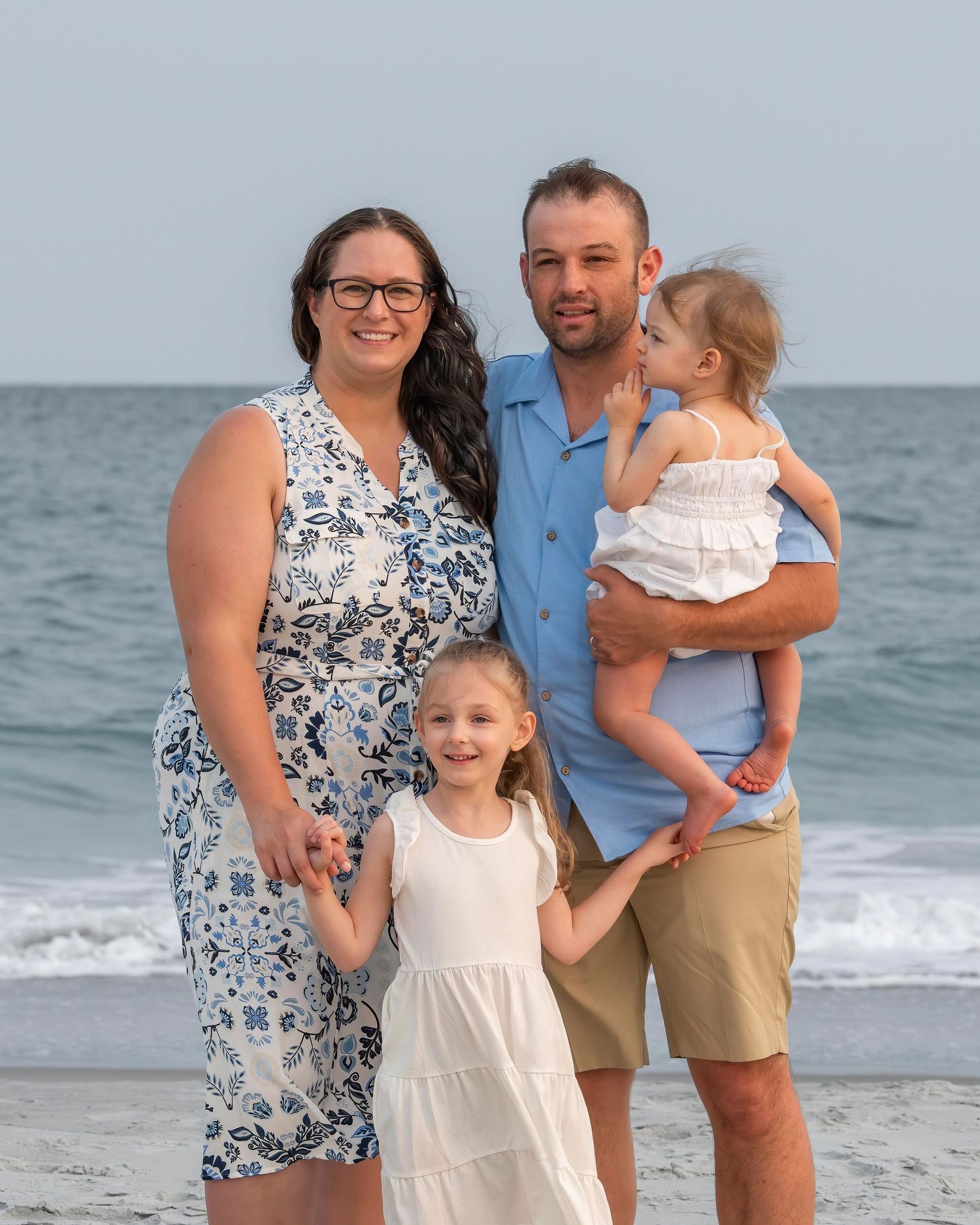 A family of four poses together on a beach with ocean waves and gray skies in the background.