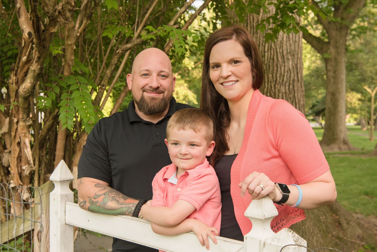 A family gathers at a white fence in a park setting with lush green trees and natural lighting in the background.