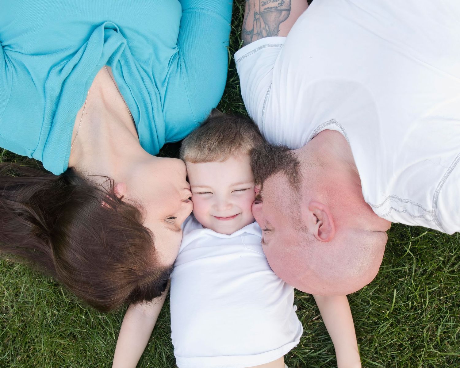 A joyful family moment captured from above as they lay together on green grass sharing kisses and smiles.