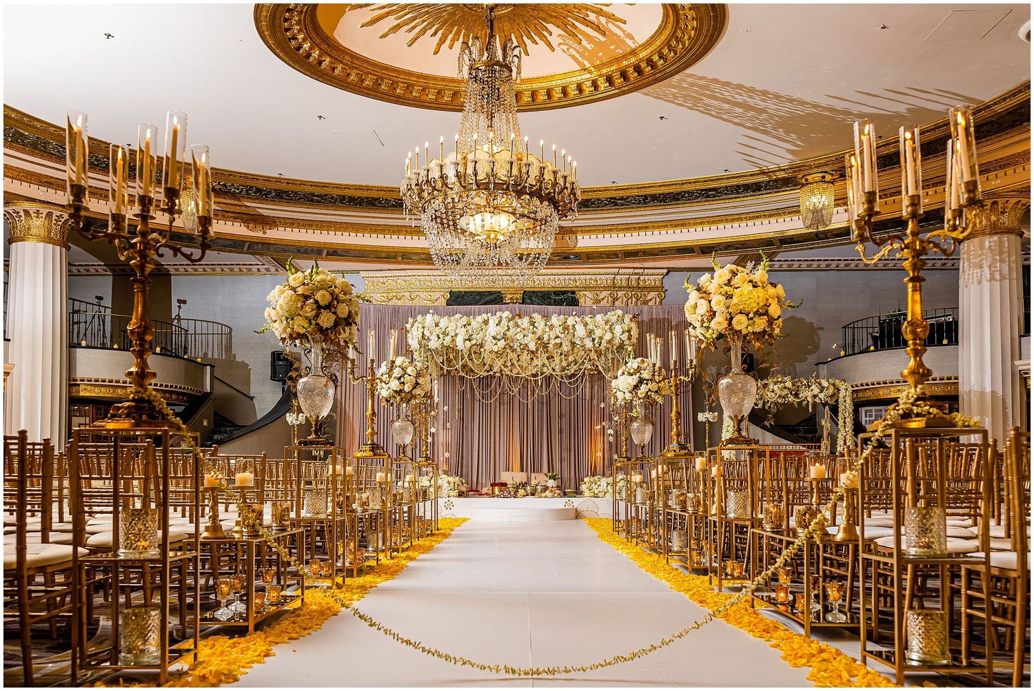 Luxurious wedding ceremony setup with gold chairs and crystal chandeliers in an ornate ballroom.