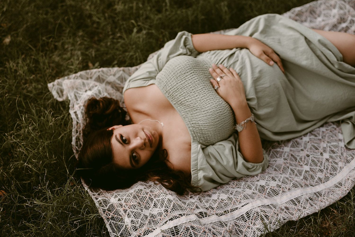 Expecting mother in grey dress rests peacefully on lace blanket outdoors.