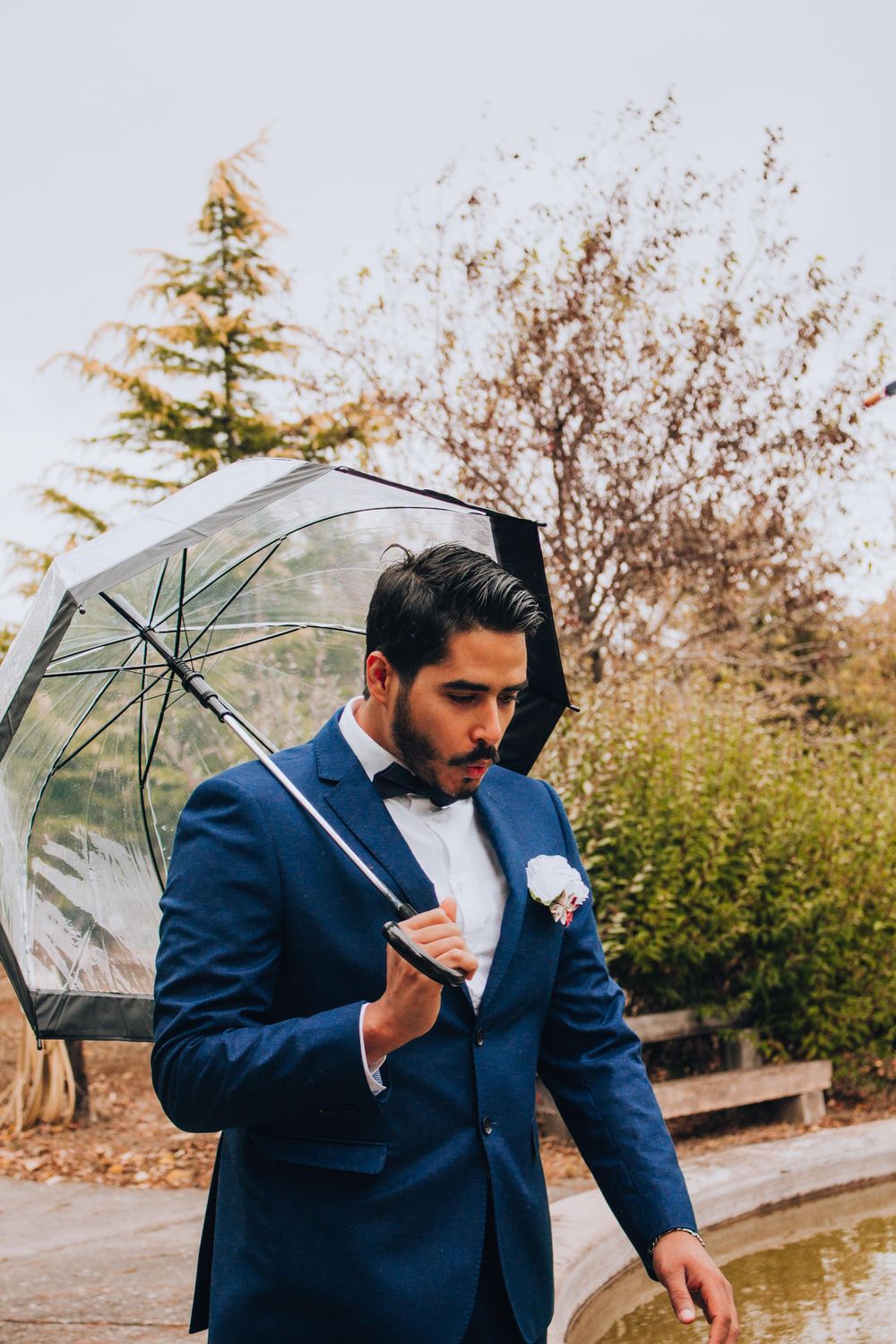 Groom in navy blue suit holding clear umbrella during autumn wedding.