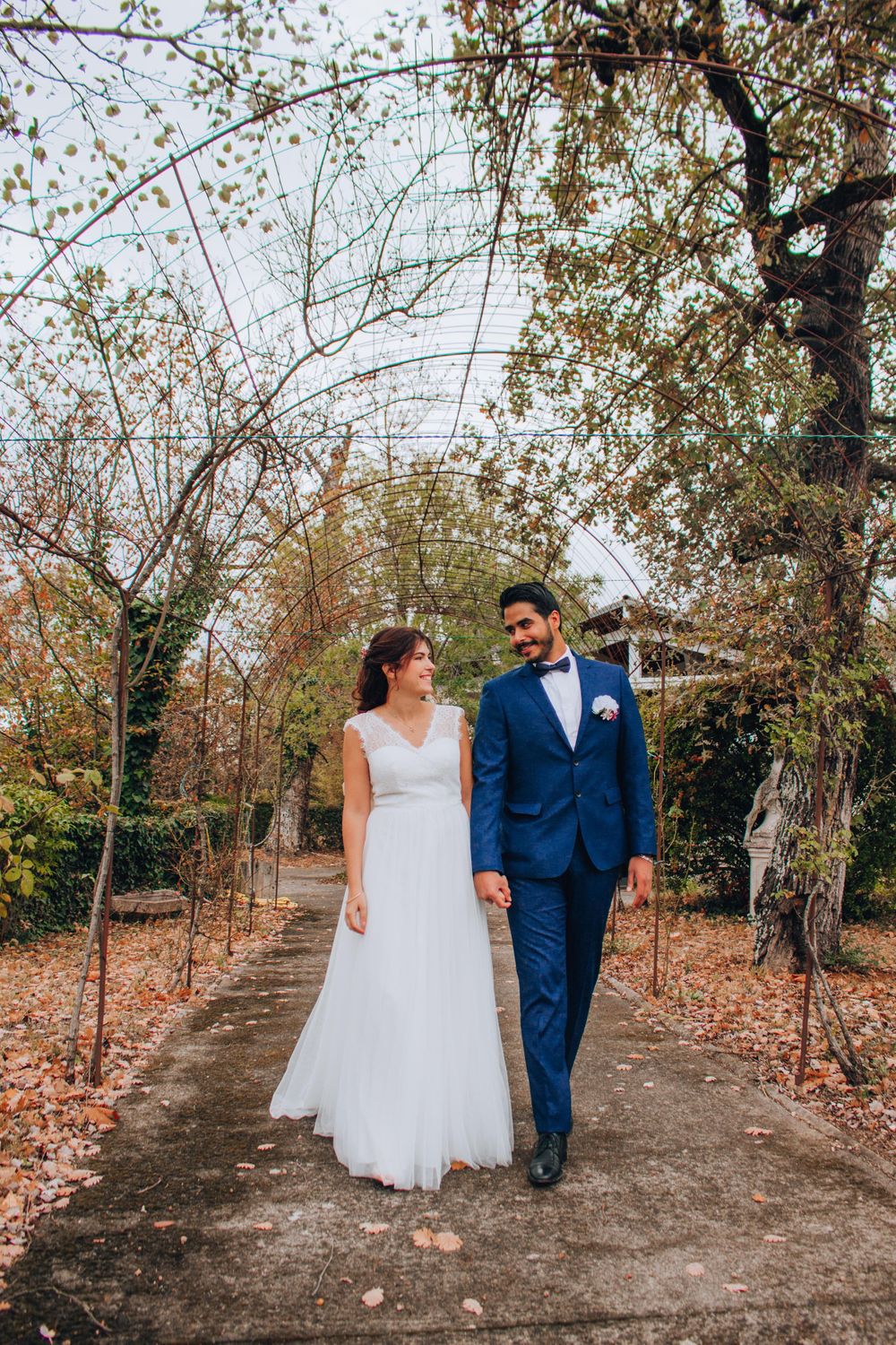 A romantic wedding couple walks down a tree-lined path during autumn, the bride in white and groom in navy blue.