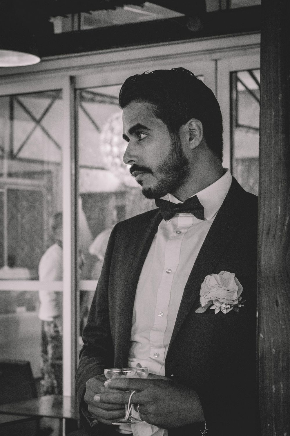 A groom in a black tuxedo with bowtie and boutonniere poses during a wedding reception.