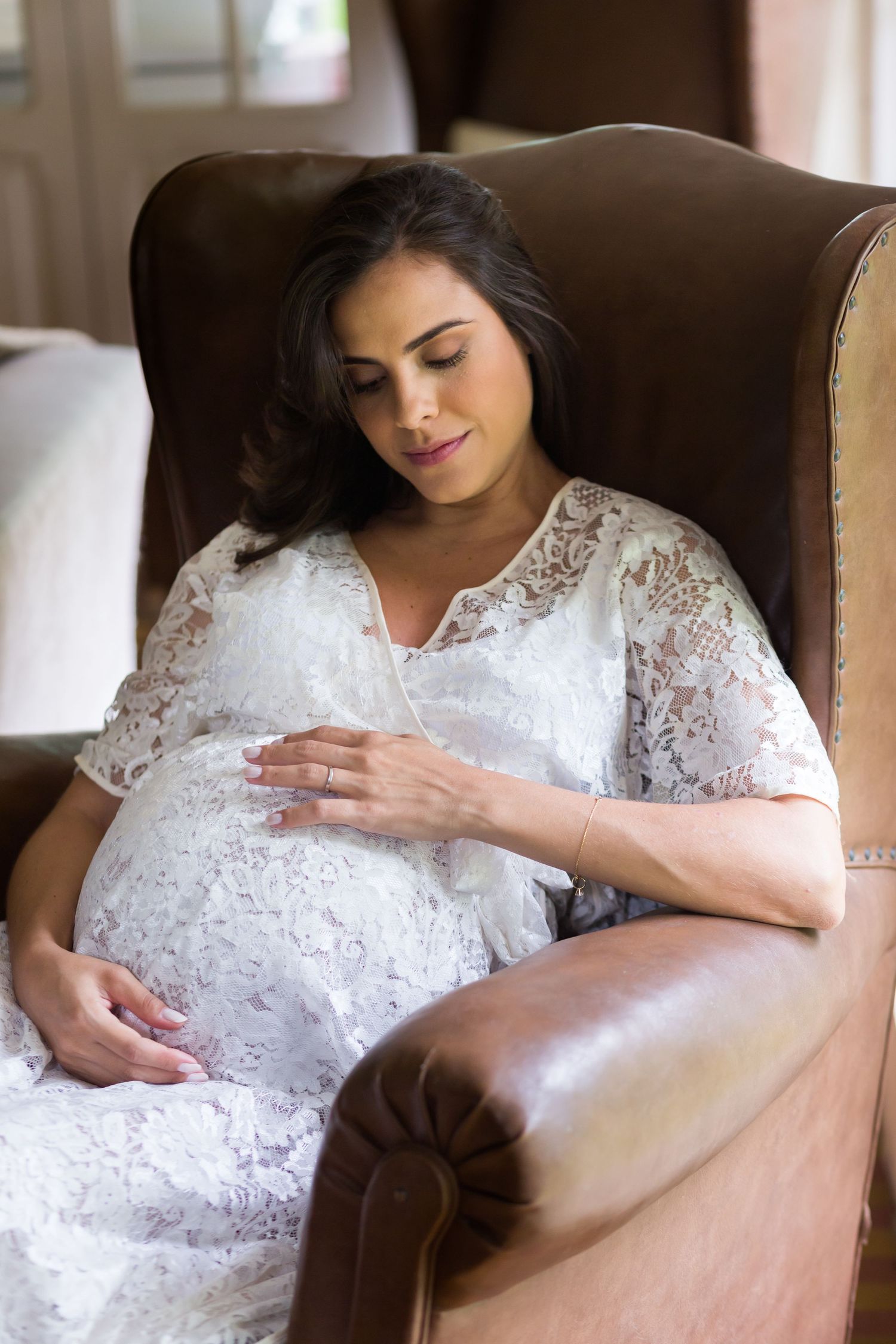 Expectant mother in white lace dress rests peacefully in leather armchair.