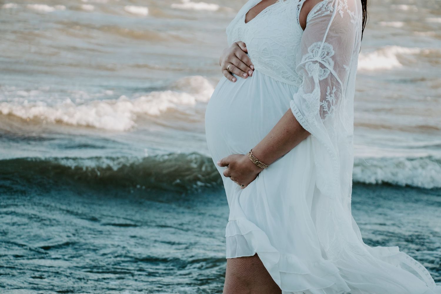 Woman in flowing white lace dress poses on beach with waves rolling in at sunset.