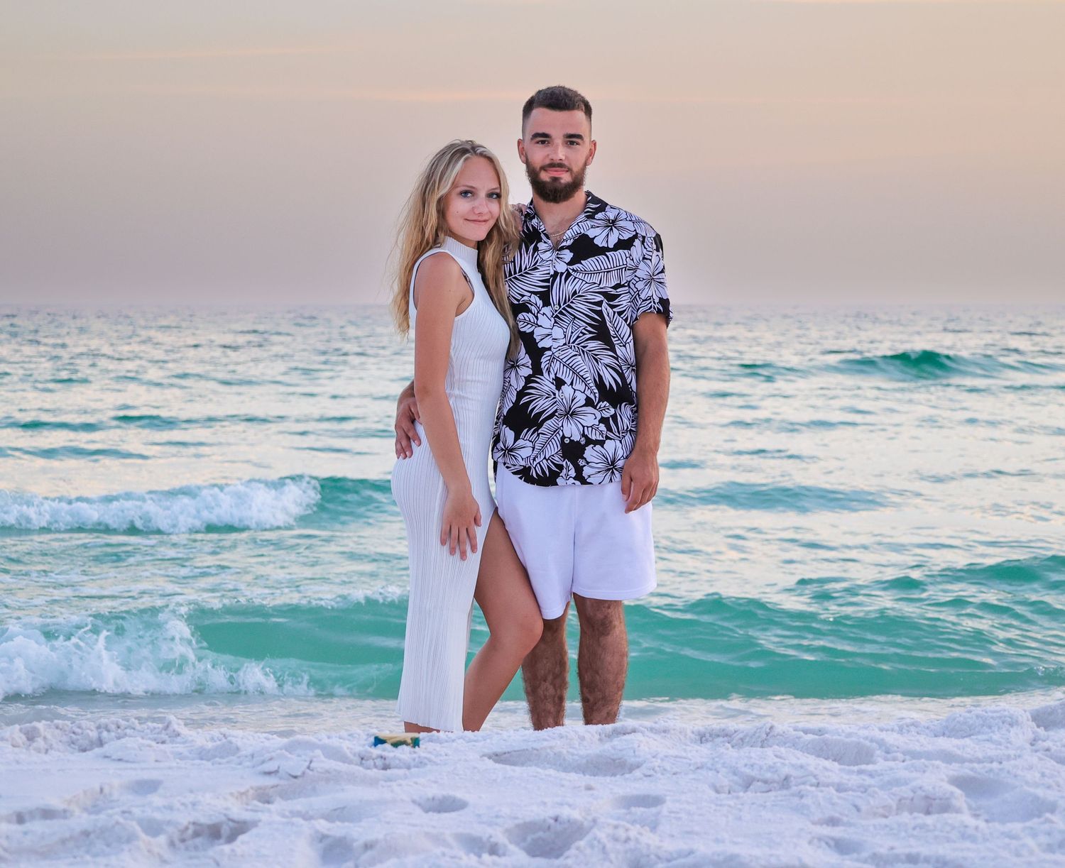 Couple in white outfits poses together on beach with turquoise waves crashing behind them.