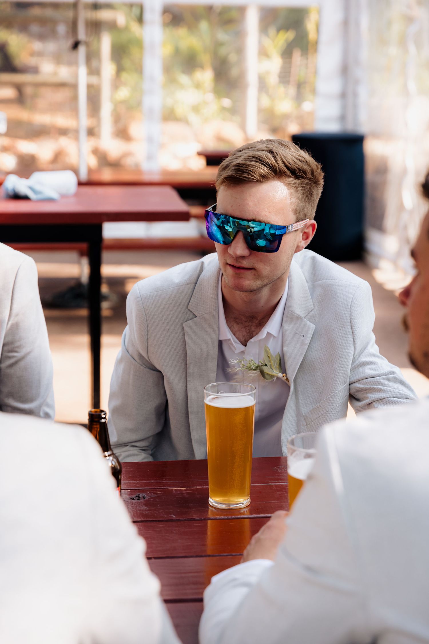 Groomsman in a light gray suit and sunglasses sitting at outdoor reception table.
