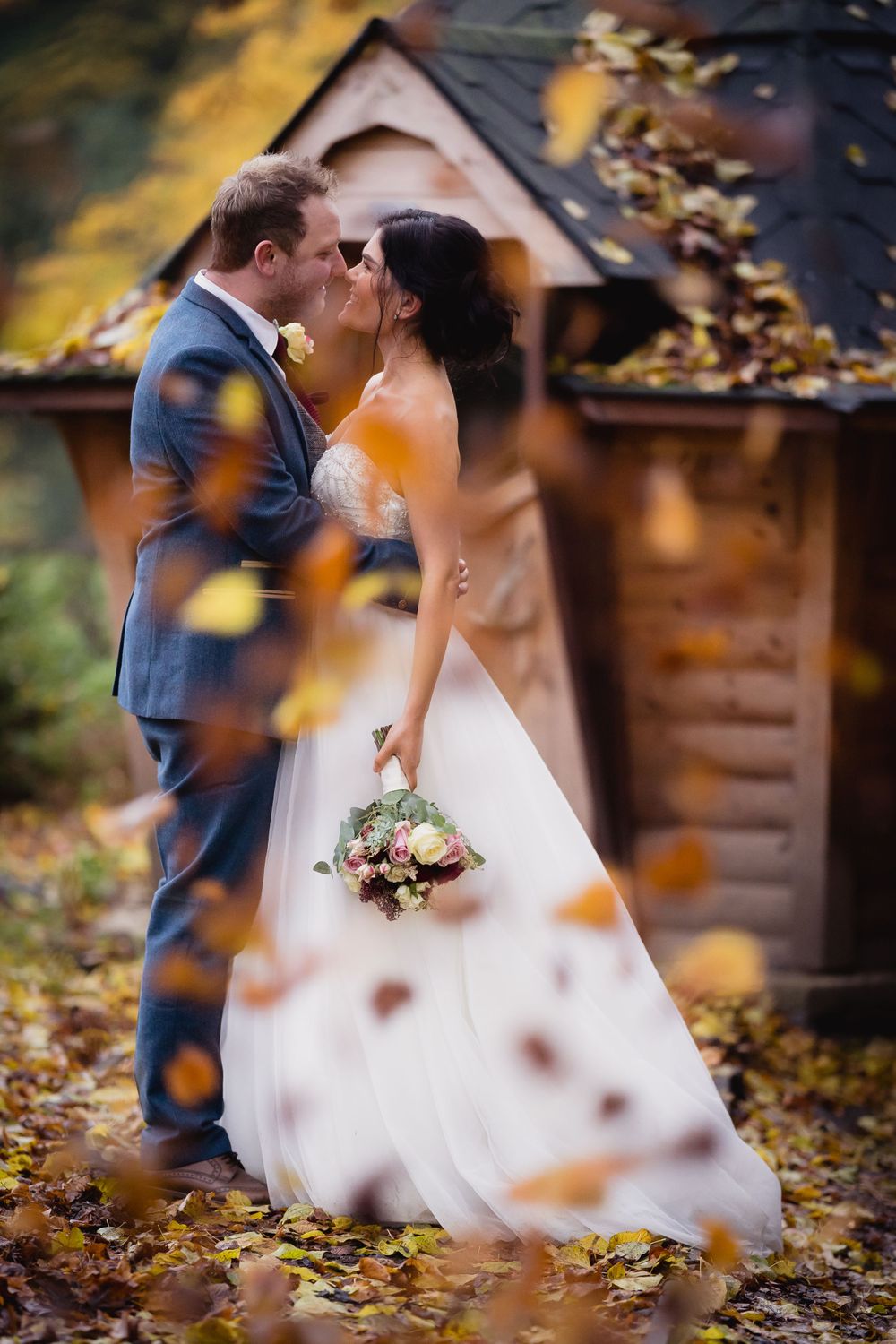 Romantic autumn wedding photo with falling leaves and rustic cabin in background.