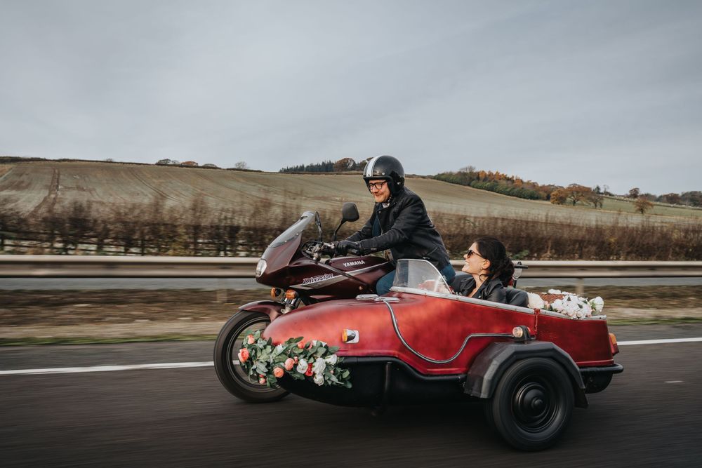 Motorcycle with vintage sidecar carries passengers along a rural road on cloudy day.