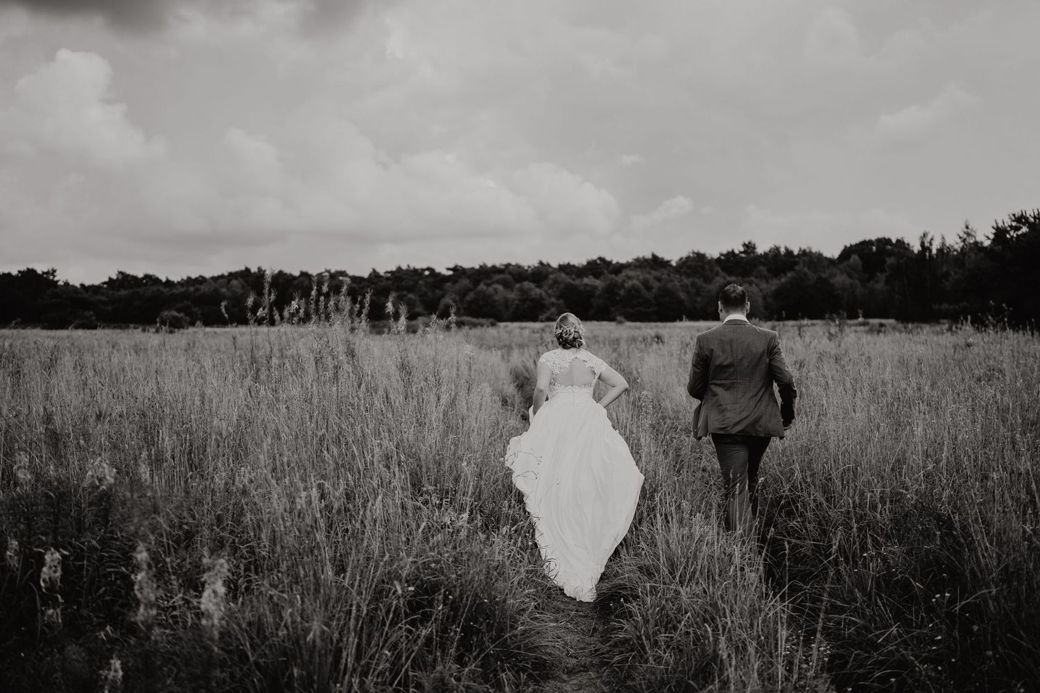 Black and white photo of newlyweds walking through rustic field on overcast day.