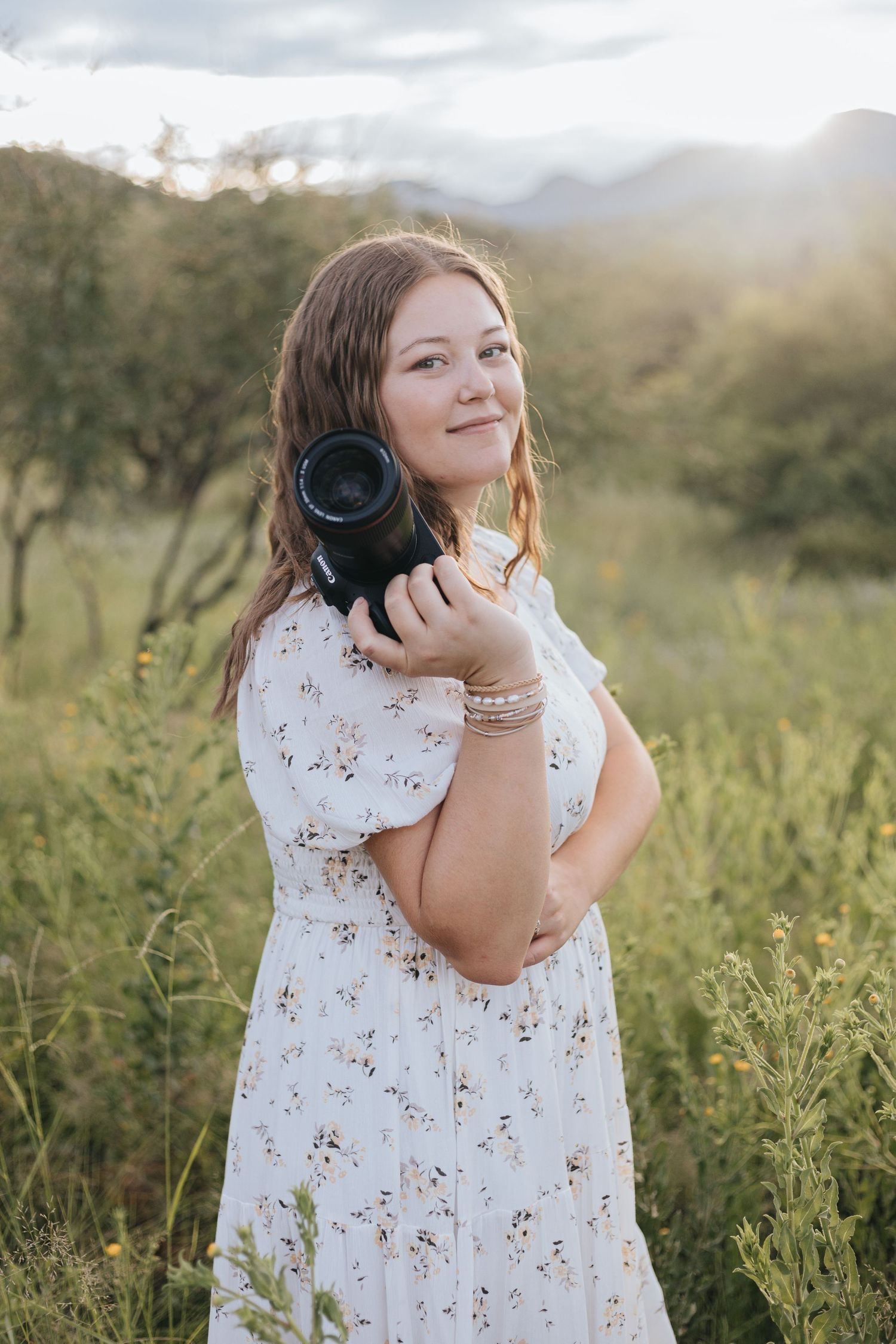 A photographer holds a professional camera in a floral dress against a natural backdrop.