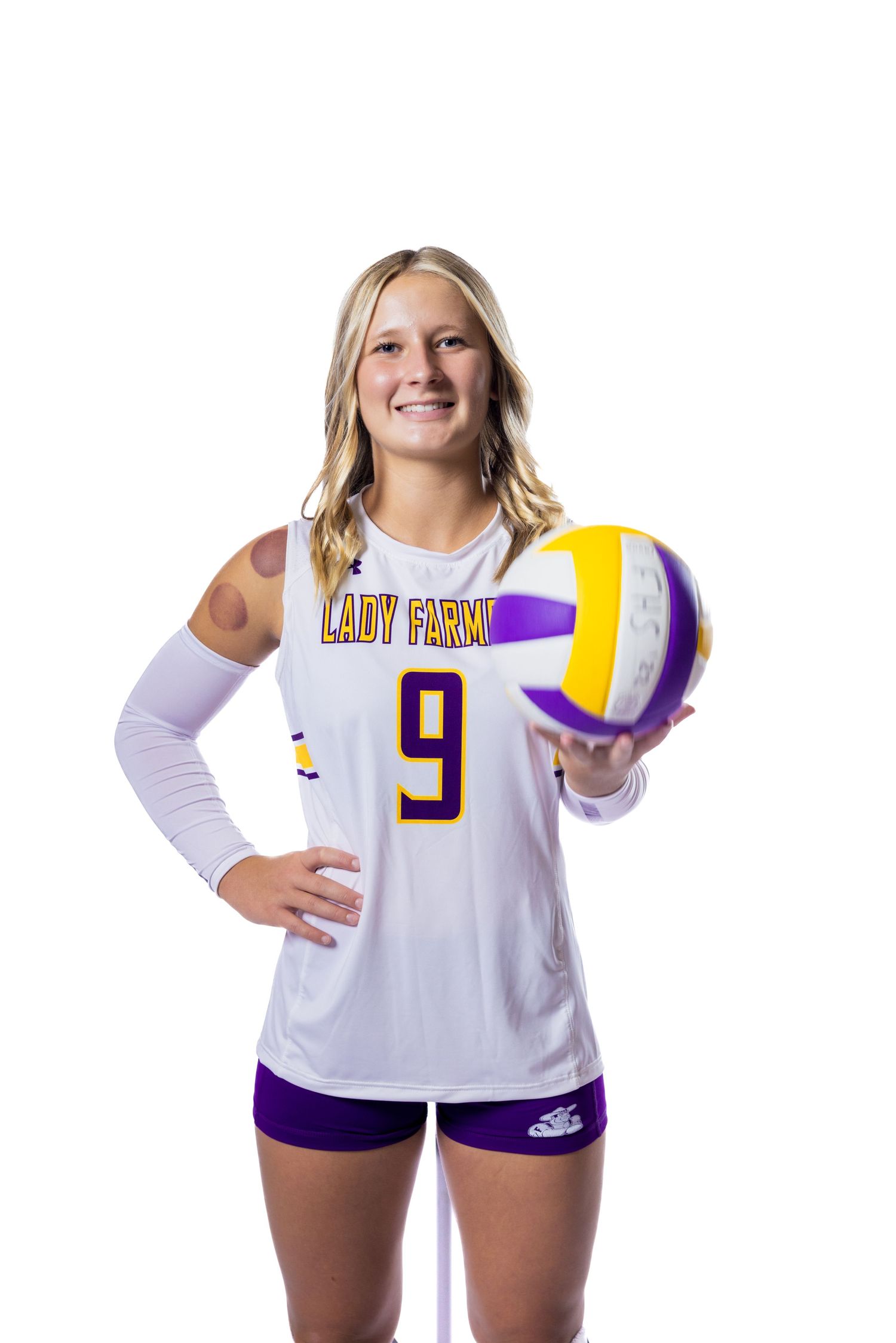 Volleyball player in white and purple uniform holds volleyball while posing for sports portrait.
