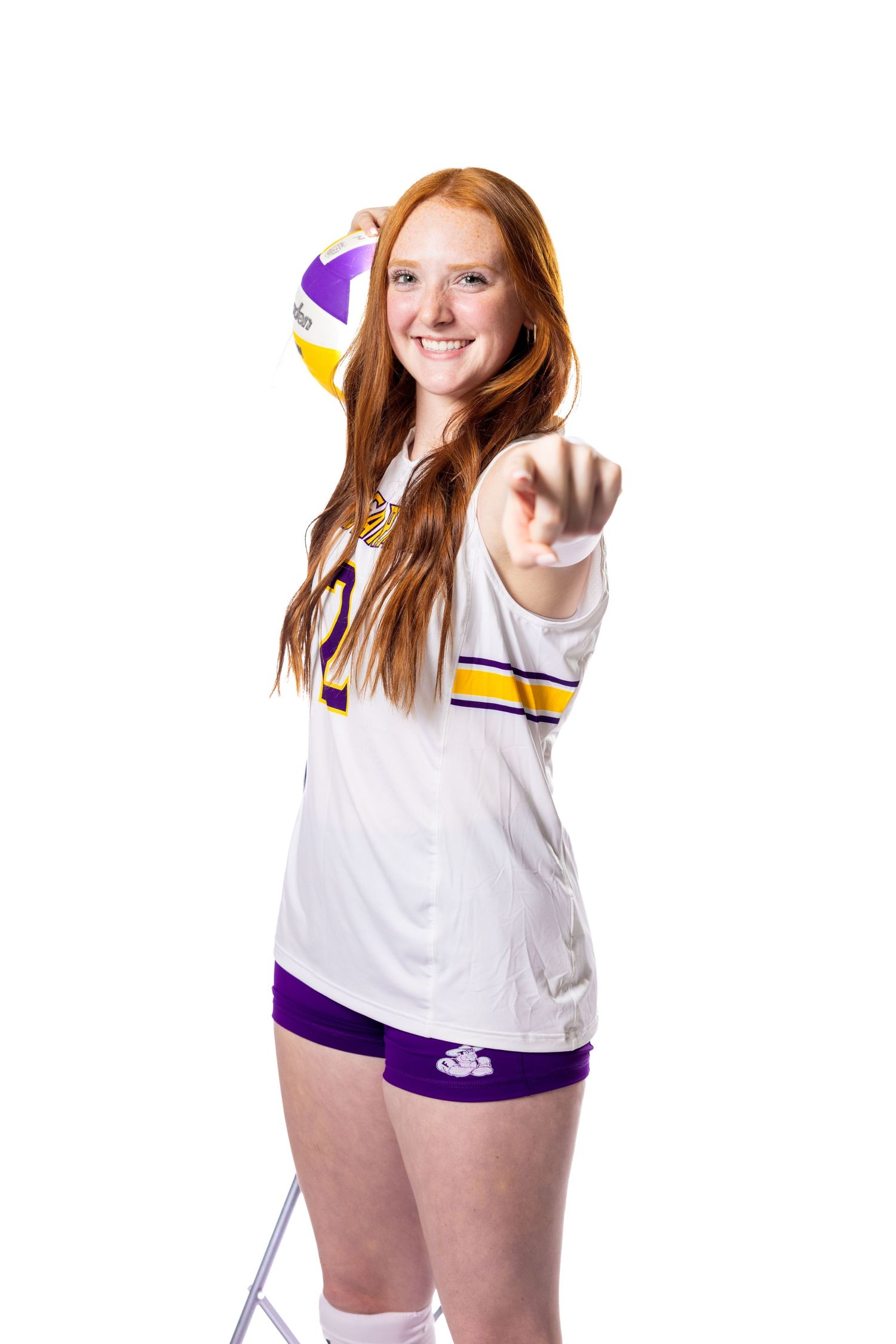 Player in white volleyball uniform points at camera while holding volleyball against white background.