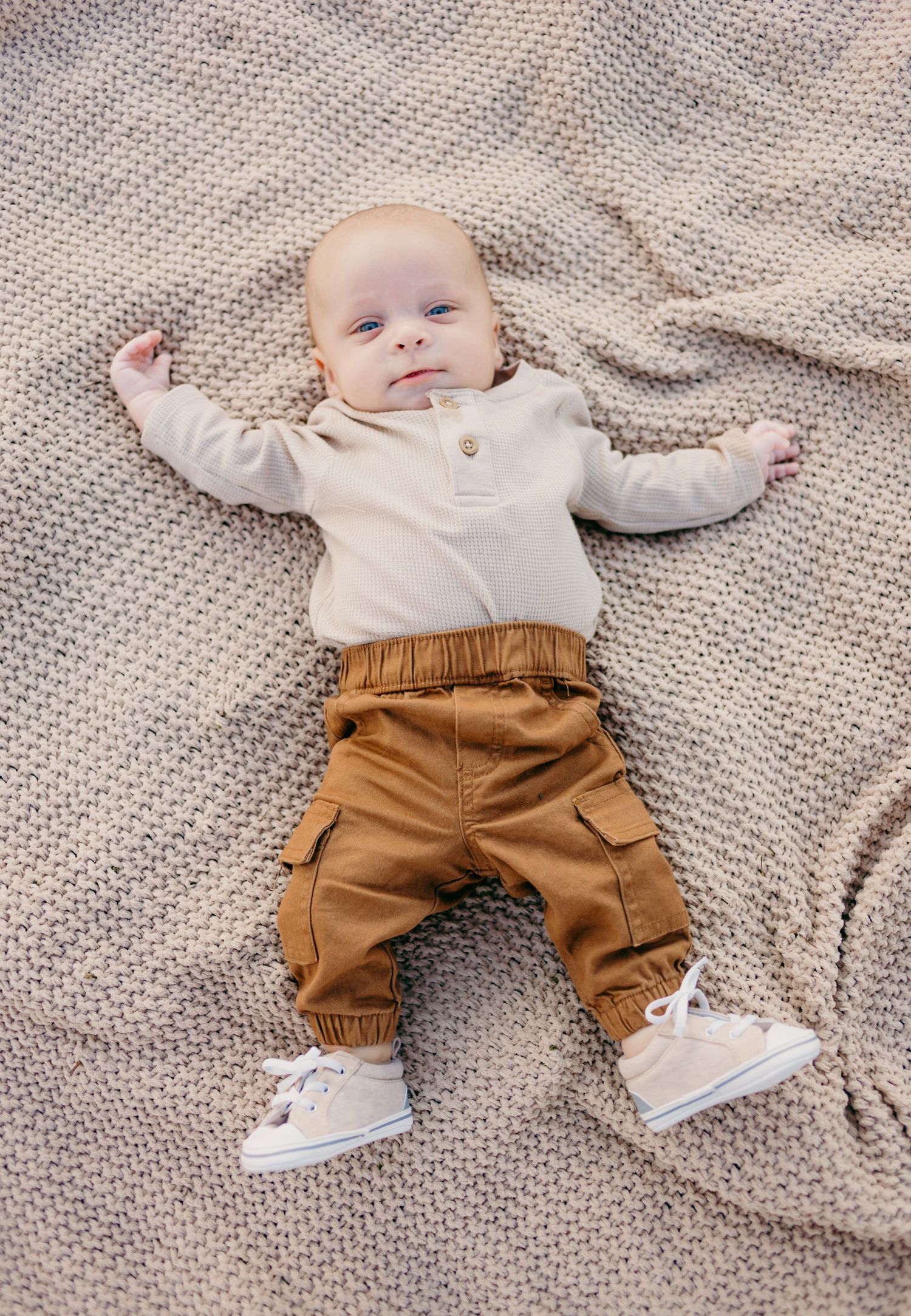Baby wearing cream sweater and brown cargo pants lies on textured gray blanket.