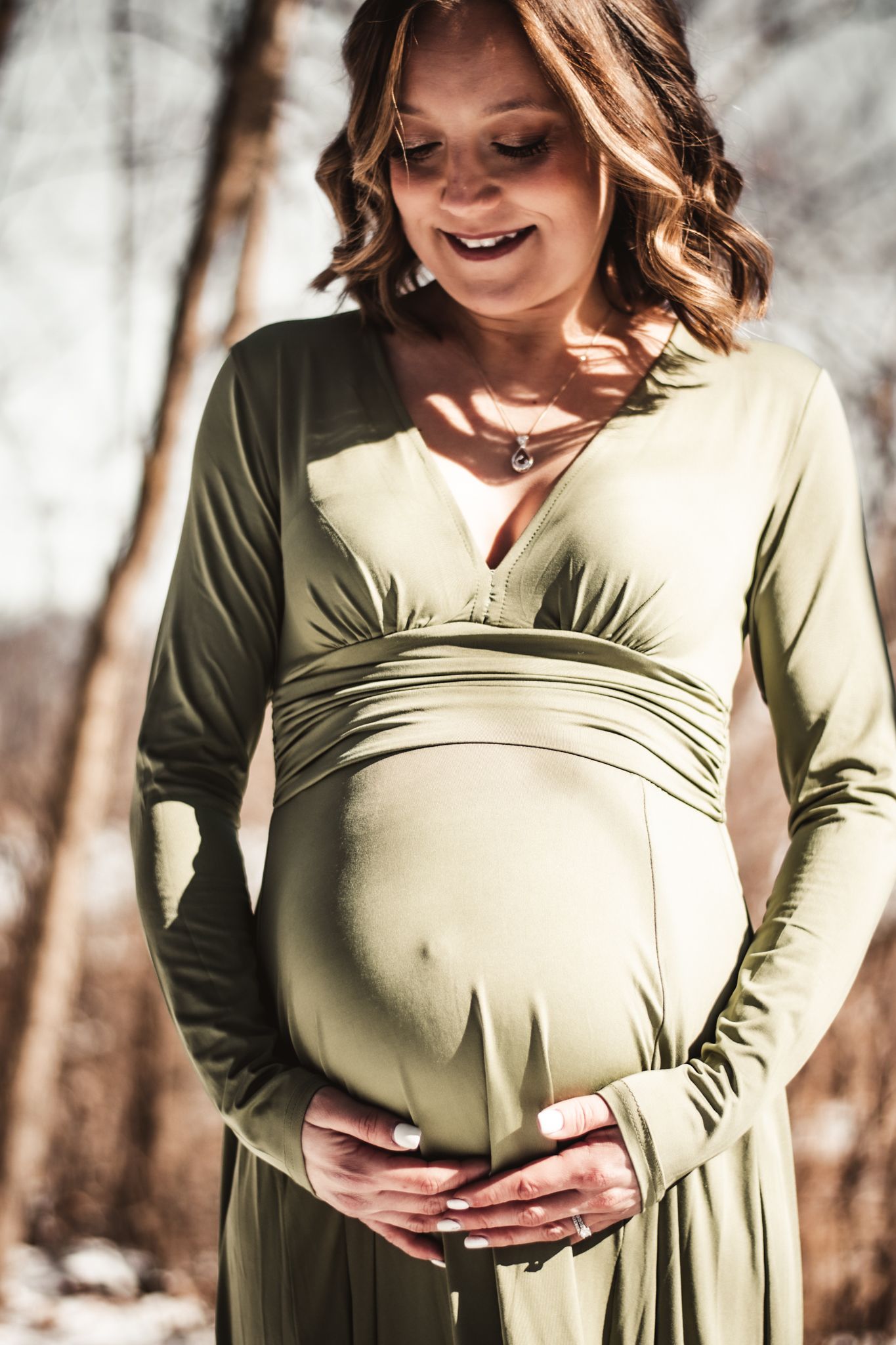 Side view of pregnant woman in sage green maternity dress smiling in winter sunlight.