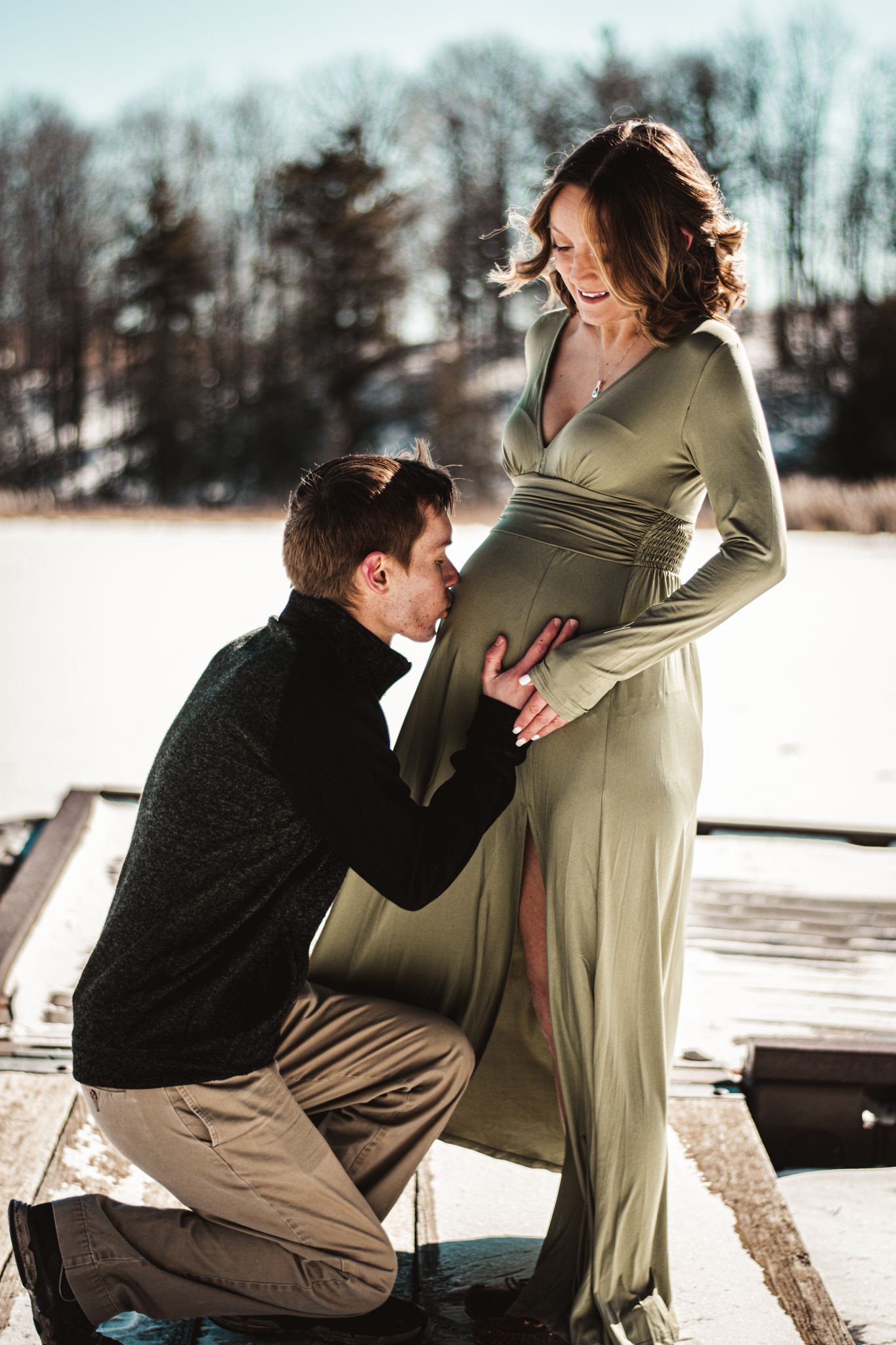 Intimate winter maternity moment captured on snowy dock with long sage dress.