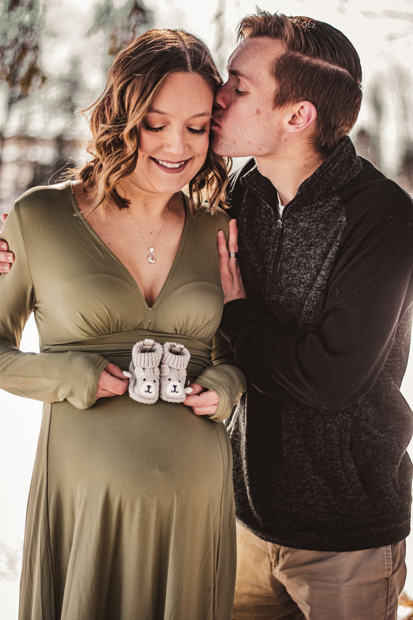 A couple shares an intimate moment while holding tiny baby shoes in a snowy winter setting.