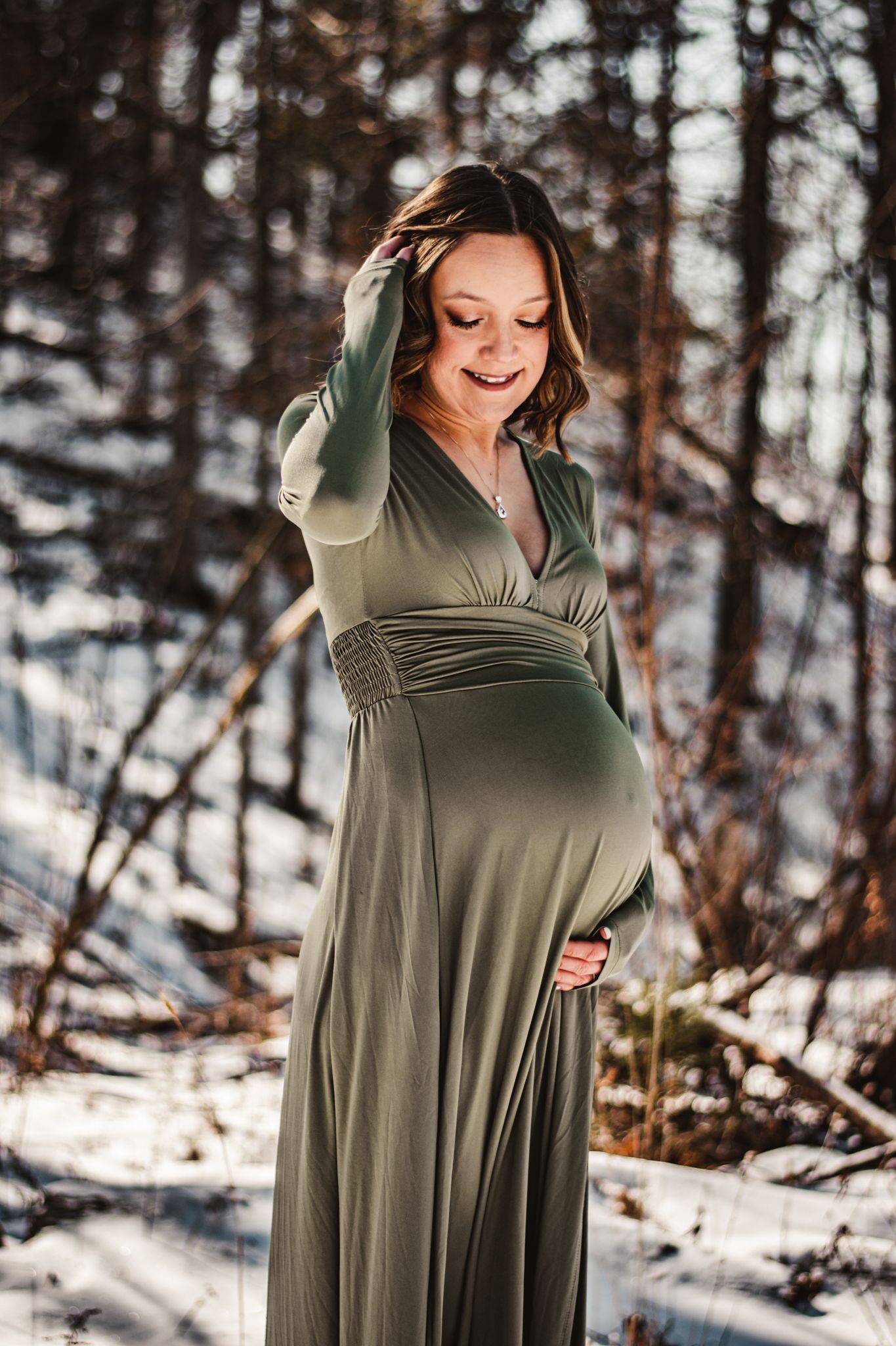 A pregnant woman in a flowing green dress touches her belly while standing in a winter forest.