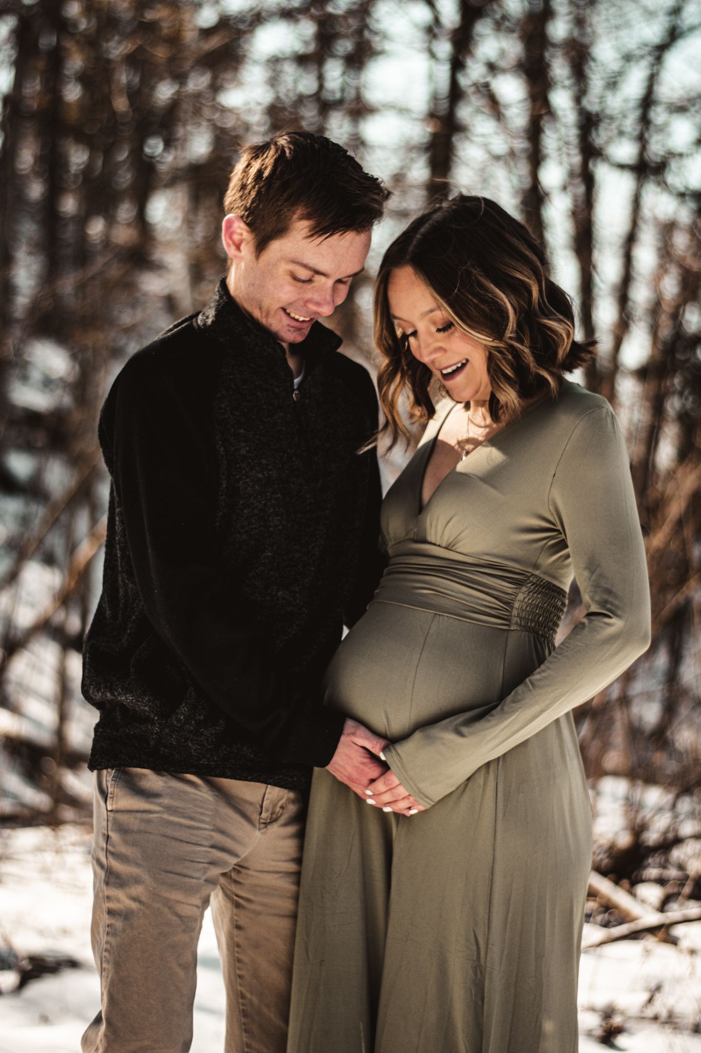 A couple shares a joyful moment during their winter maternity photoshoot in a forest setting.
