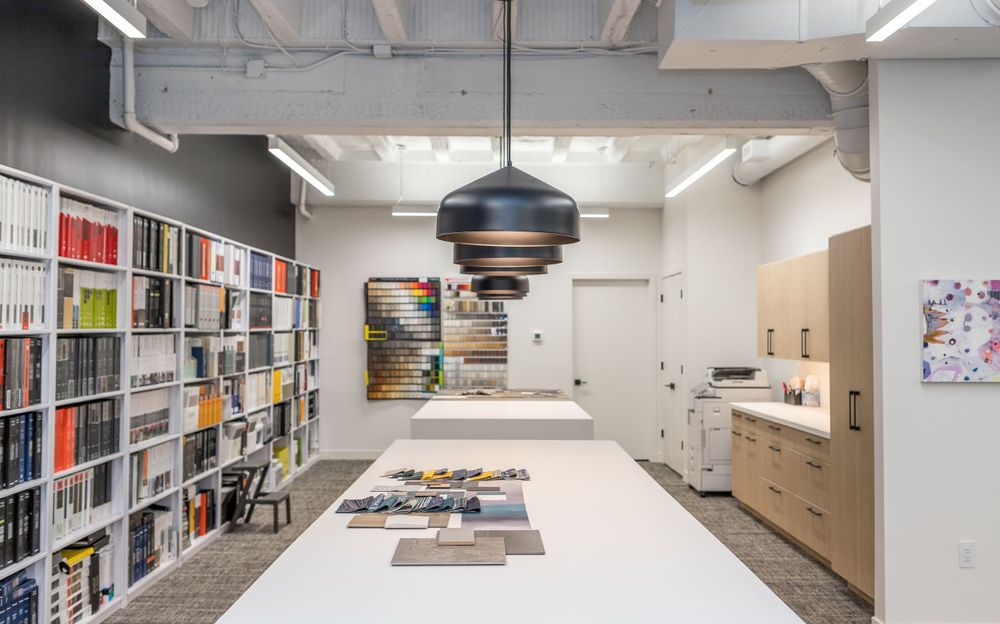 Modern office workspace with large bookshelf and central work table under pendant lighting.
