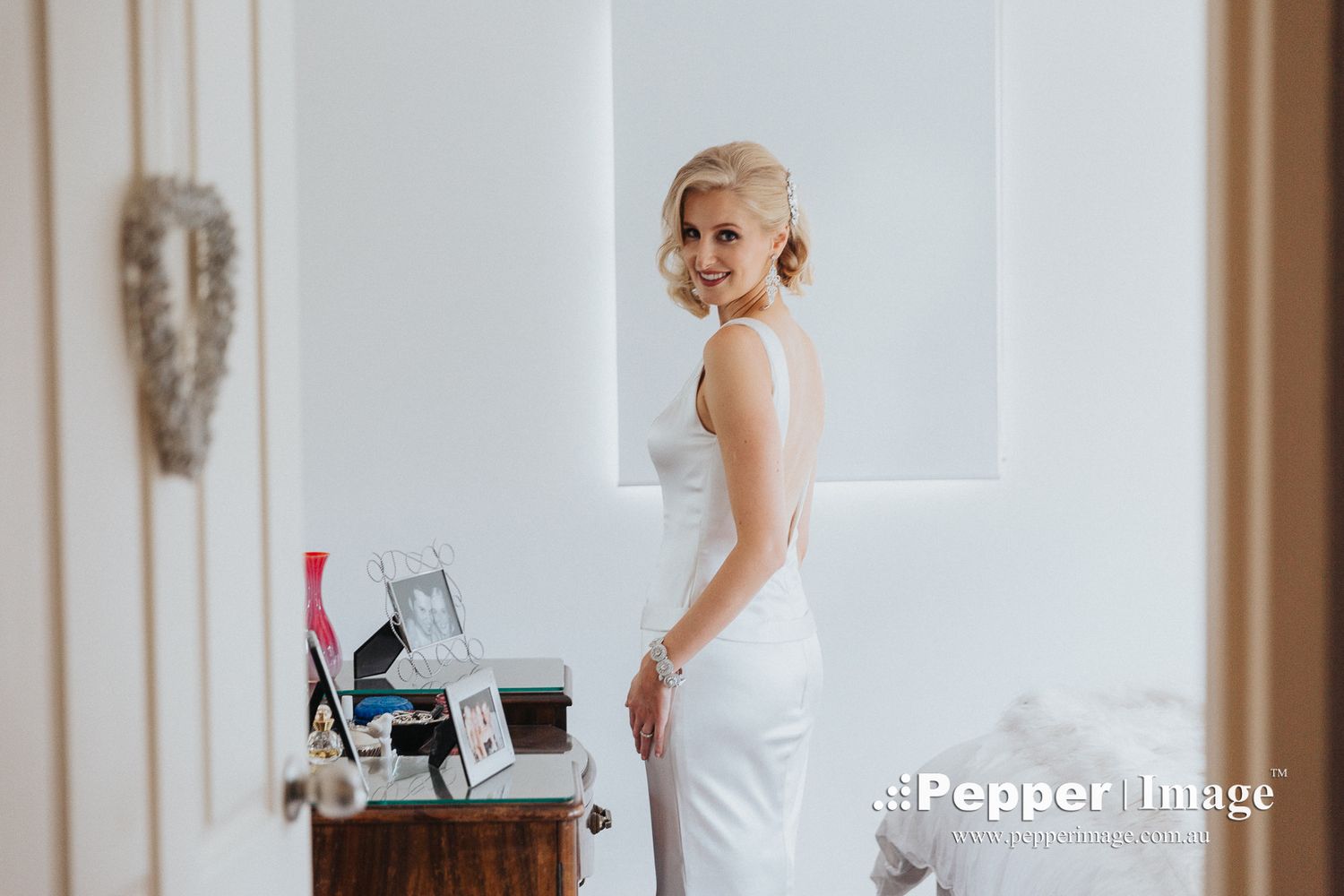 Smiling blonde bride in a sleek white wedding dress posing in a bright white room before ceremony.