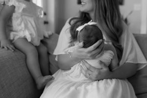 black and white image. detail shot of moms hands gently holding her newborn with her toddlers feet resting on her legs