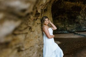 A senior in a white dress leaned against the rock of the inside of a cave in the mountains