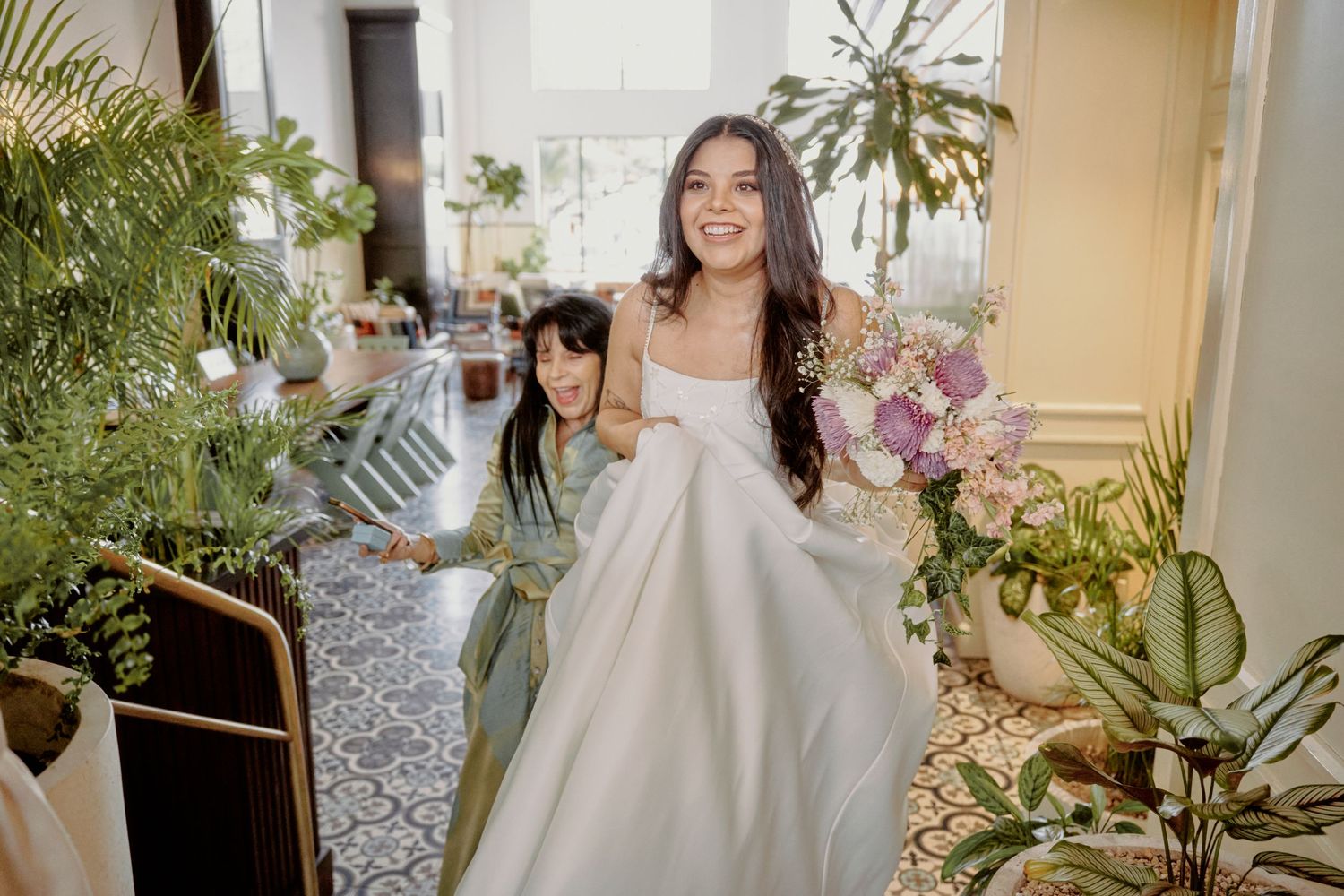 A bride in a flowing white gown walks through a tropical plant-filled venue.