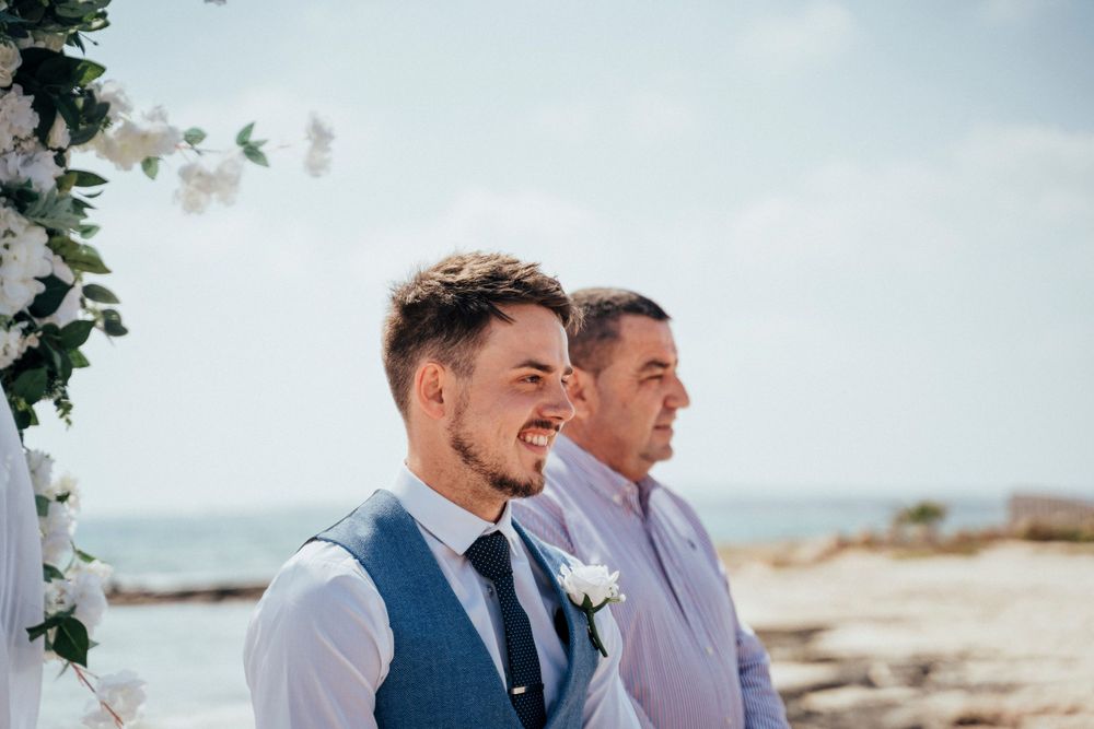 Two wedding attendees stand near floral arch with ocean view in background.