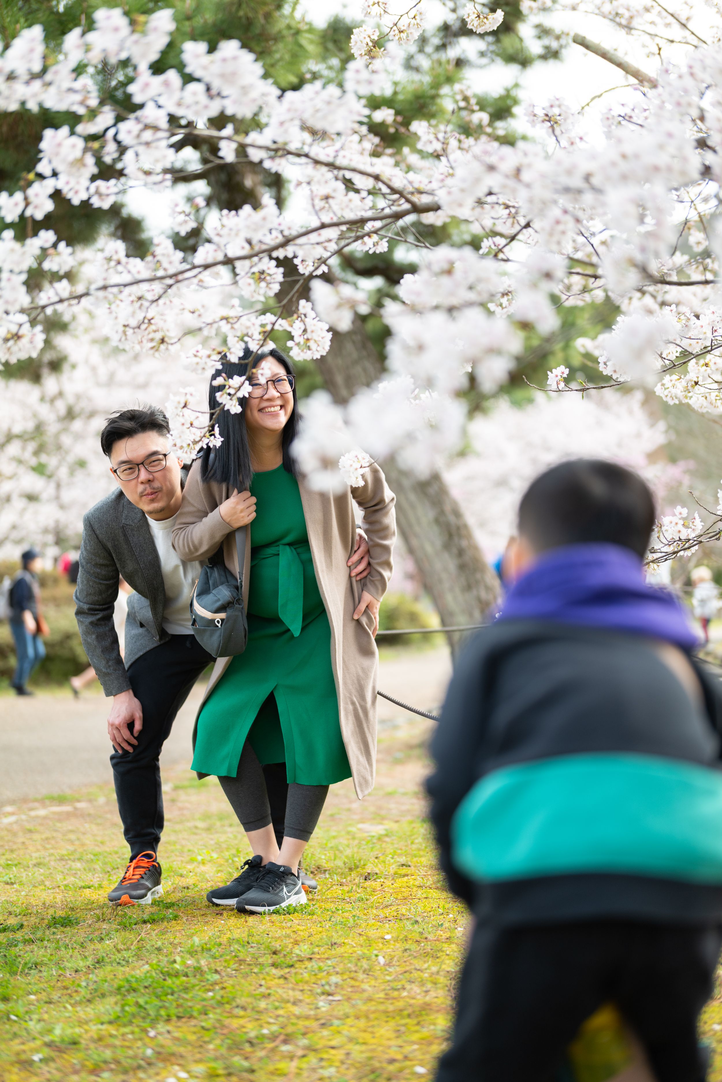 Asian couple posing under cherry blossom tree while child takes their photo outdoors.