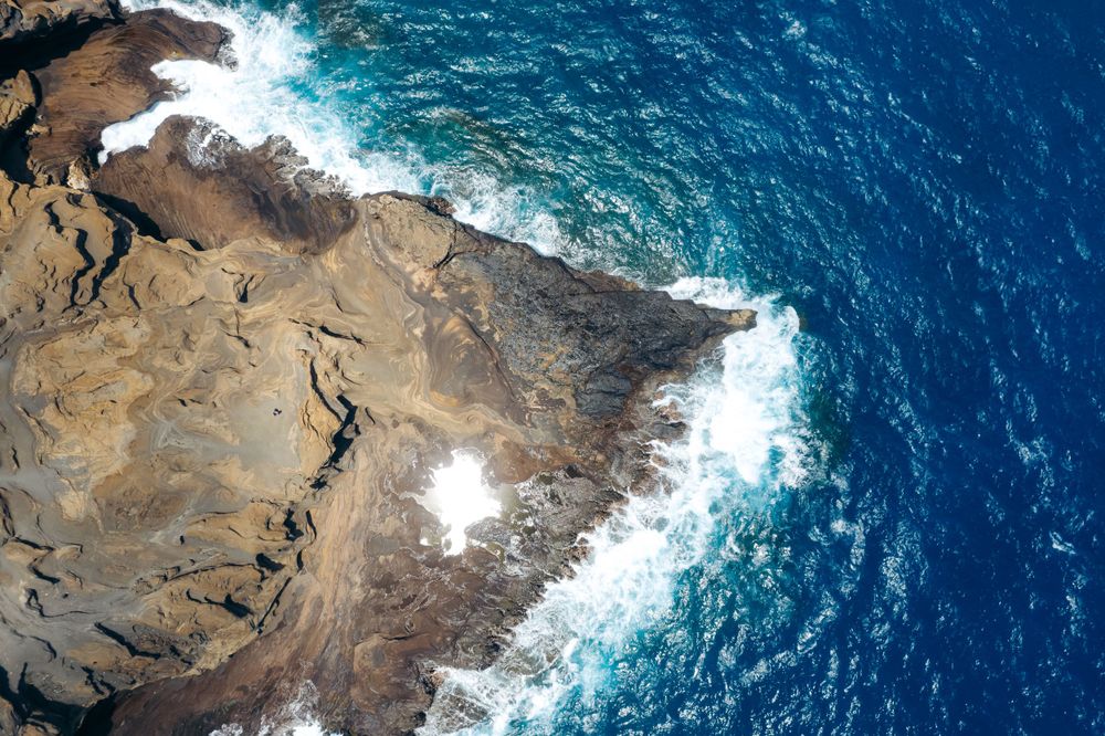 Rocky coastal cliffs meet deep blue ocean waters with white waves crashing against the shore.