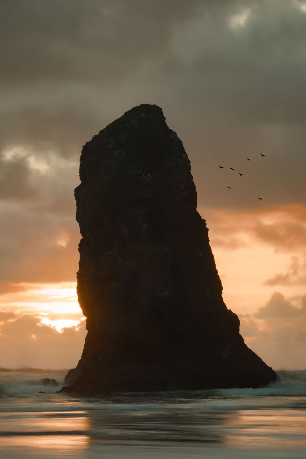 Towering sea stack silhouetted against orange sunset sky with birds flying.