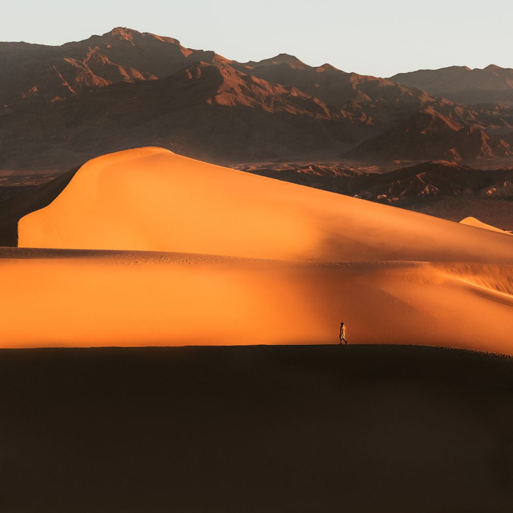 A tiny silhouette stands on golden sand dunes against a backdrop of rugged mountains at sunset.