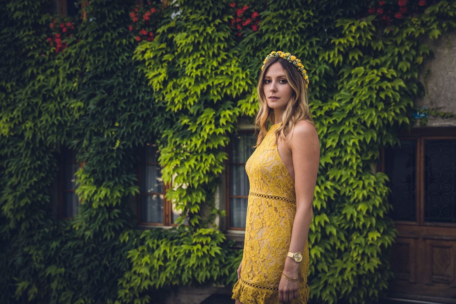 Woman in yellow lace dress poses against ivy-covered wall in garden setting.