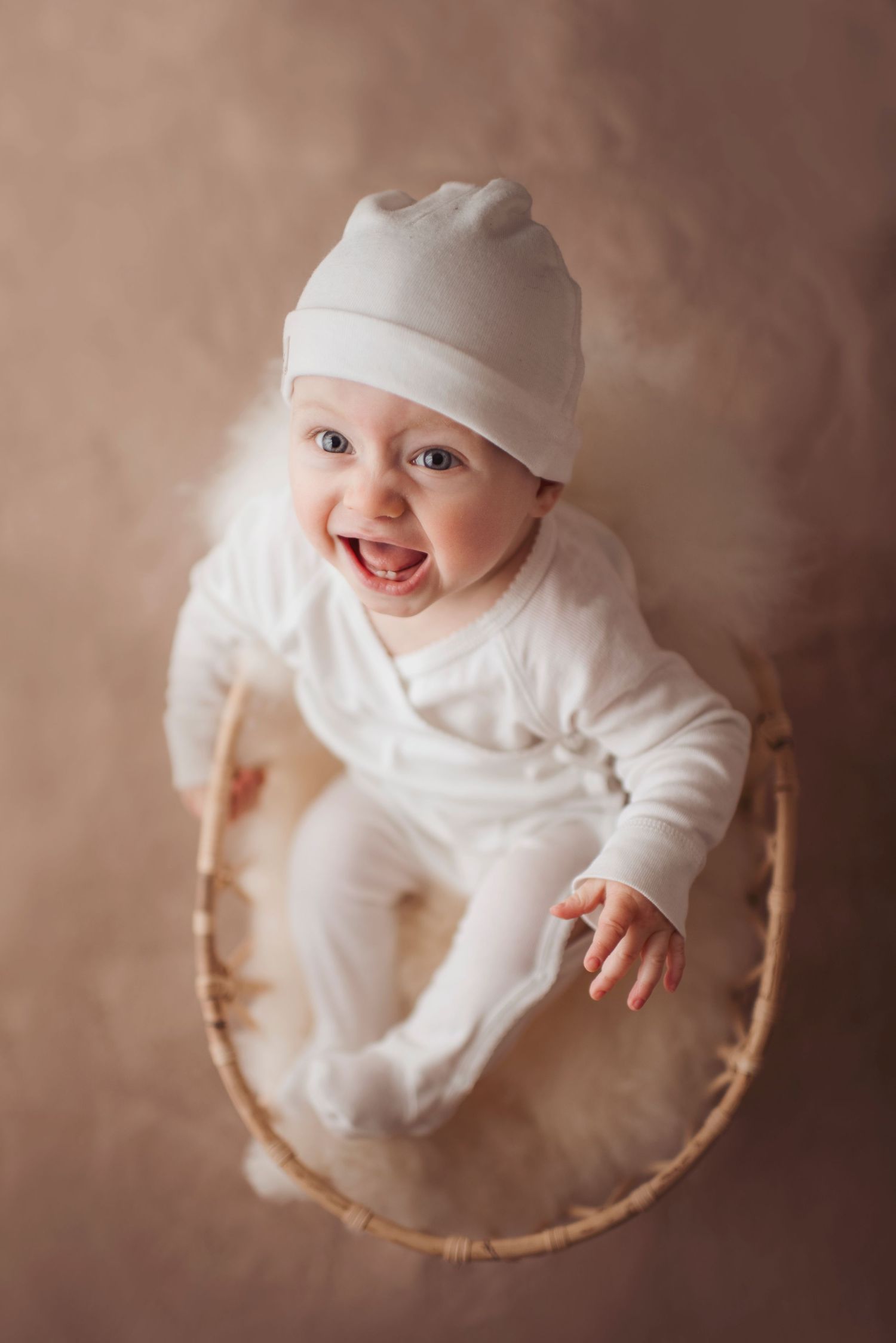 Happy baby in white outfit and cap sitting in woven basket during studio photoshoot.