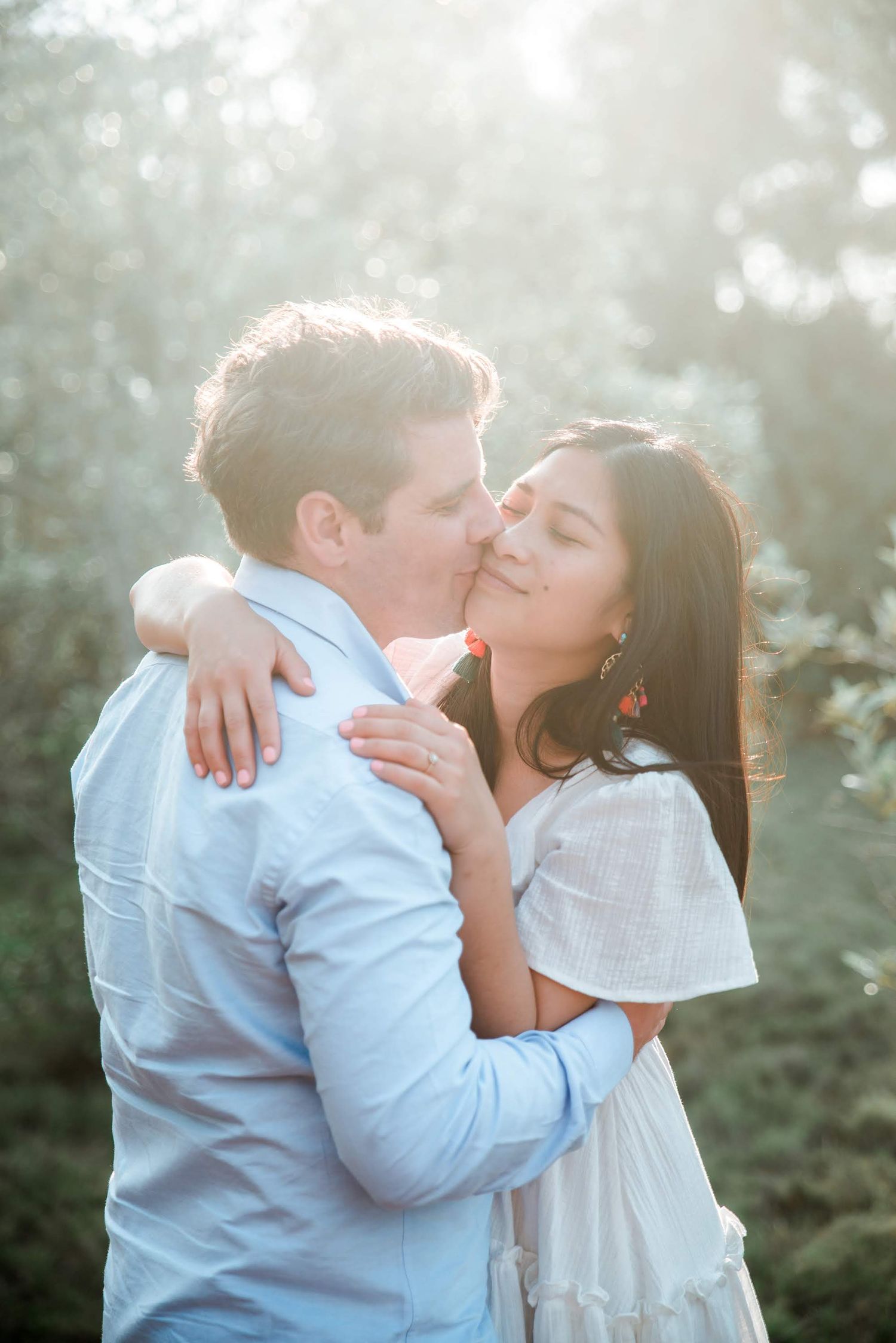 An intimate couple embraces in soft sunlit forest setting wearing light colored clothing.
