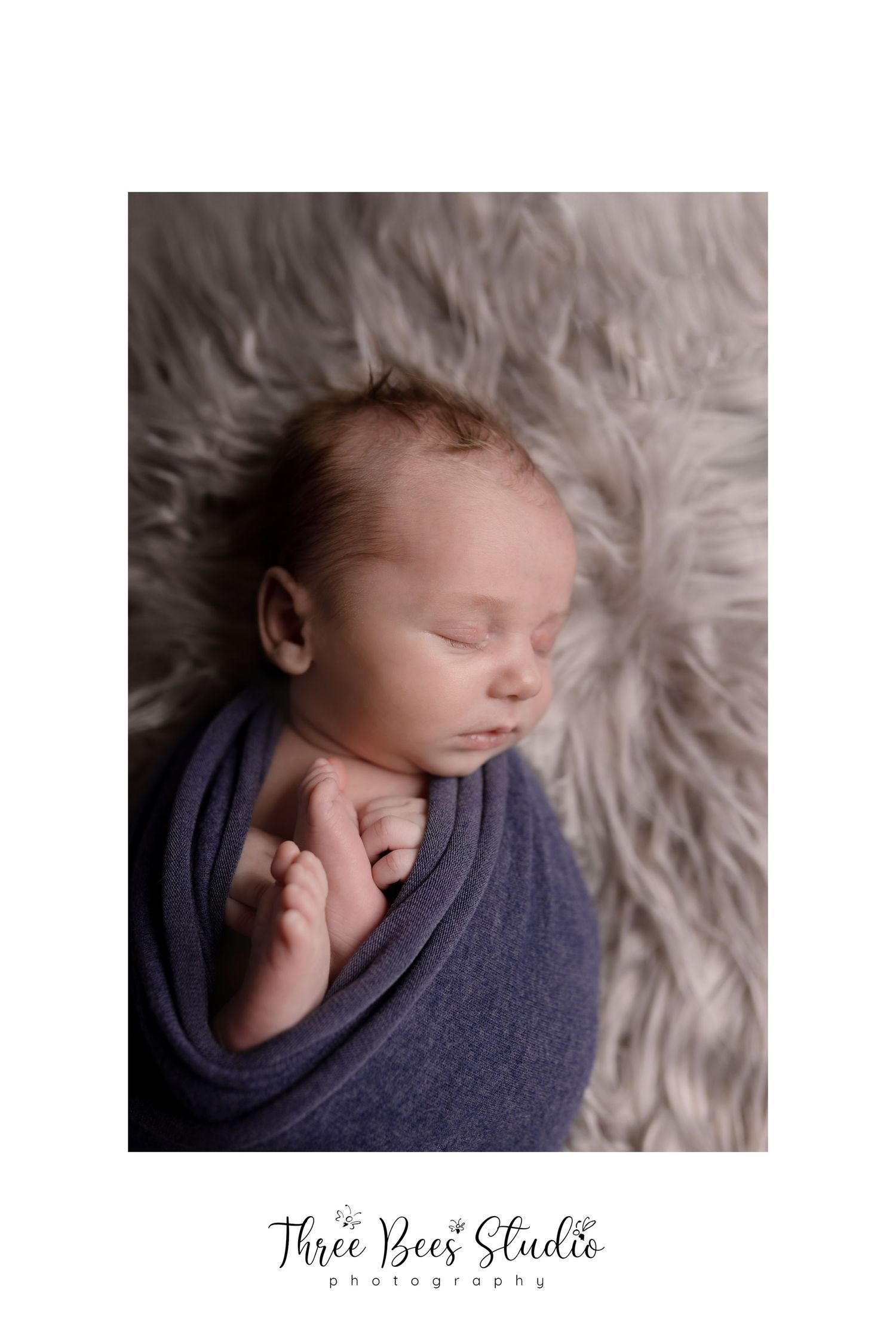 Sleeping newborn wrapped in navy blue swaddle on soft grey fur blanket.