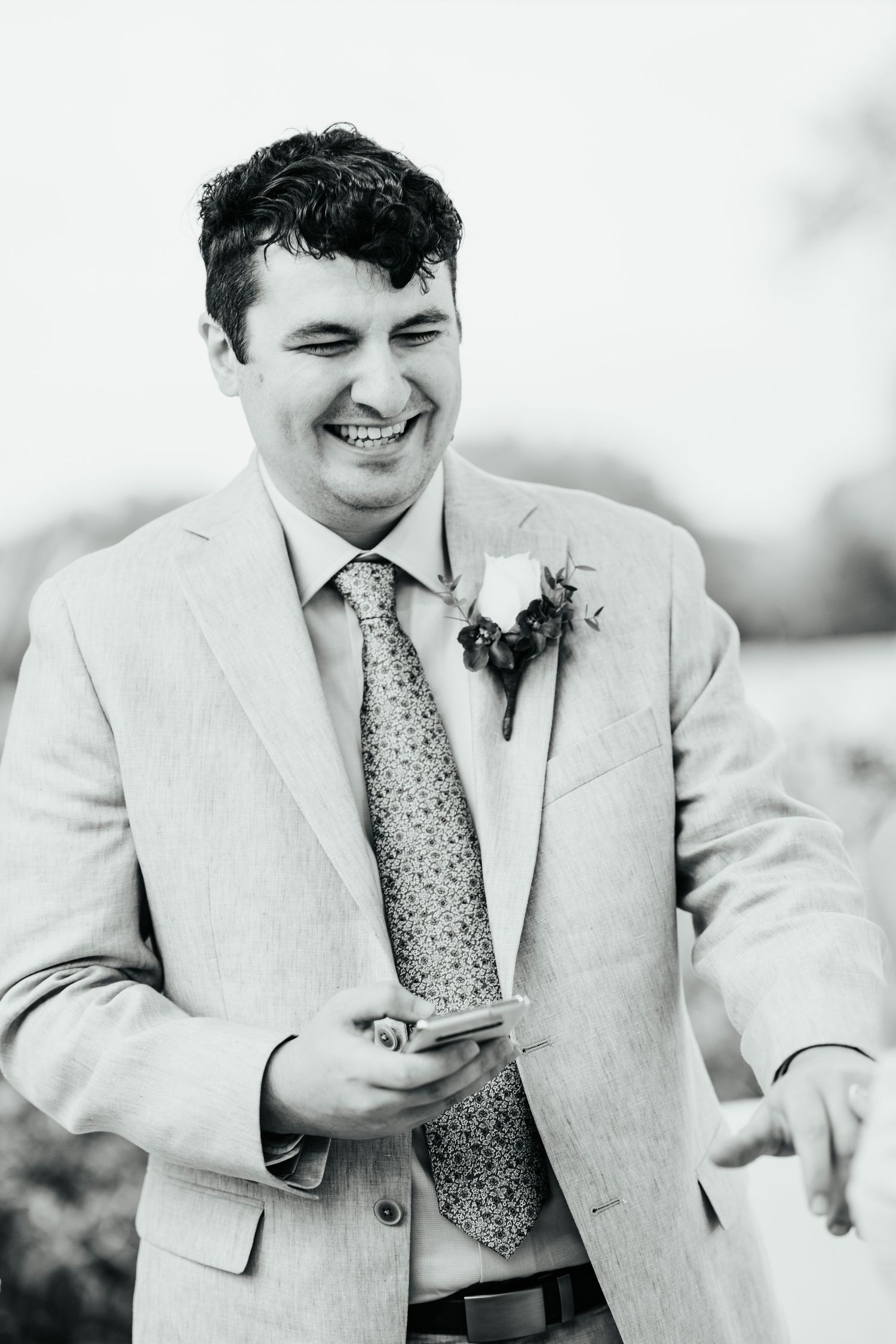 Groom in light beige suit laughing while looking at phone during wedding.