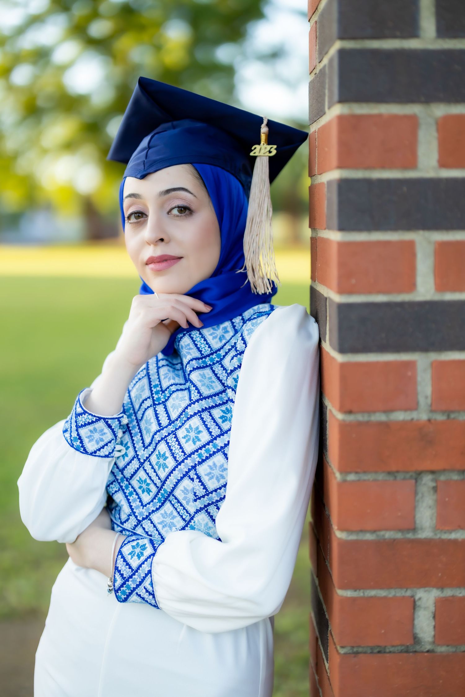 Graduate in blue cap and gown poses against brick wall outdoors.