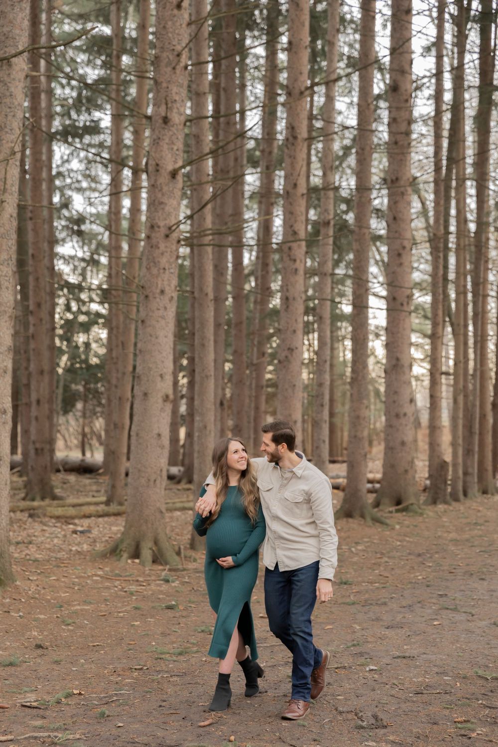 pregnant couple walking through the woods and smiling at each other at Stoney Creek
