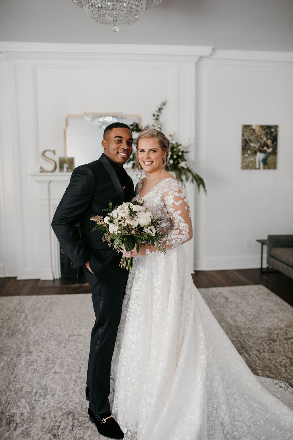 Smiling interracial wedding couple posing indoors, bride holding white floral bouquet in lace gown.