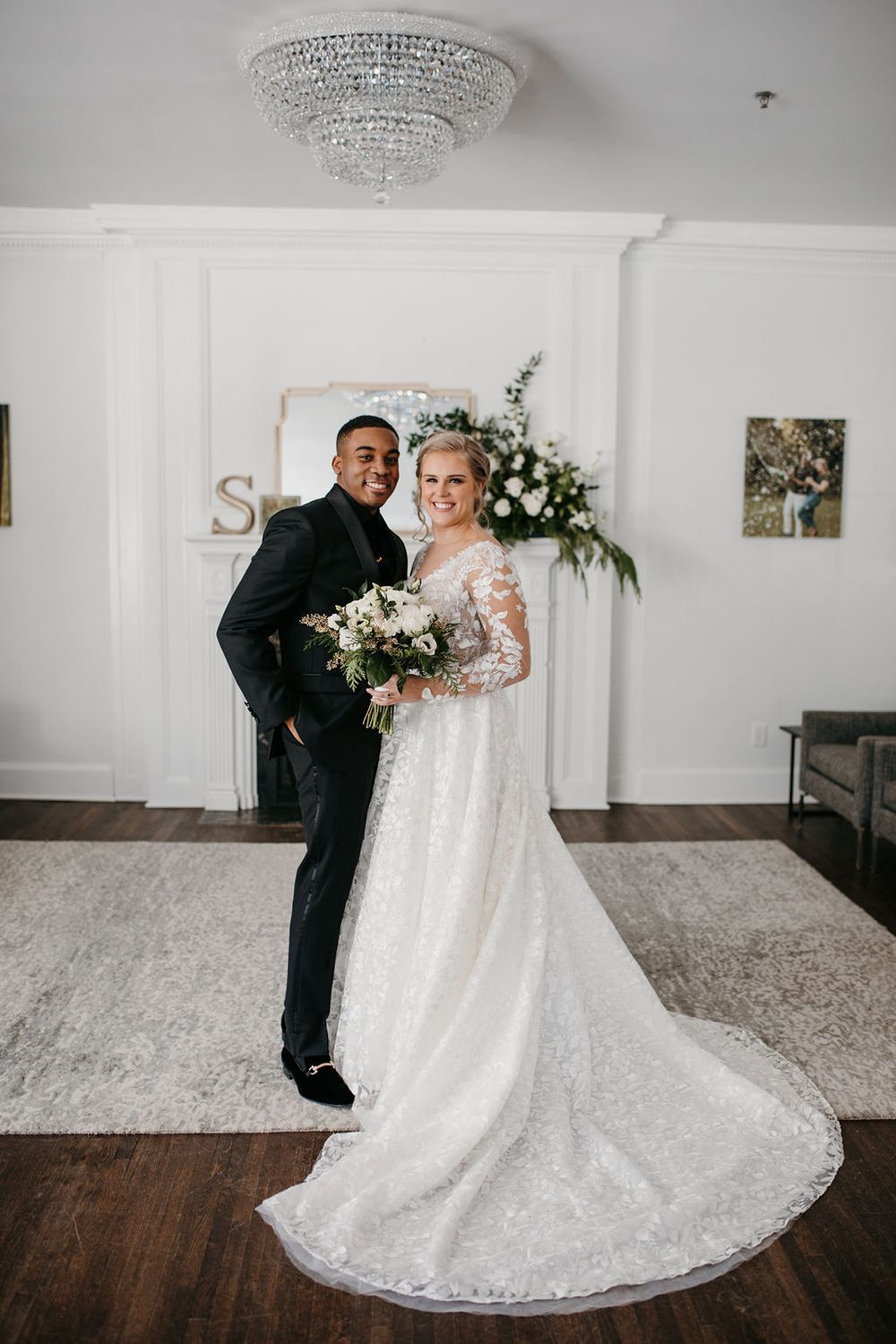 Bride and groom posing under crystal chandelier, bride in lace gown holding white bouquet.