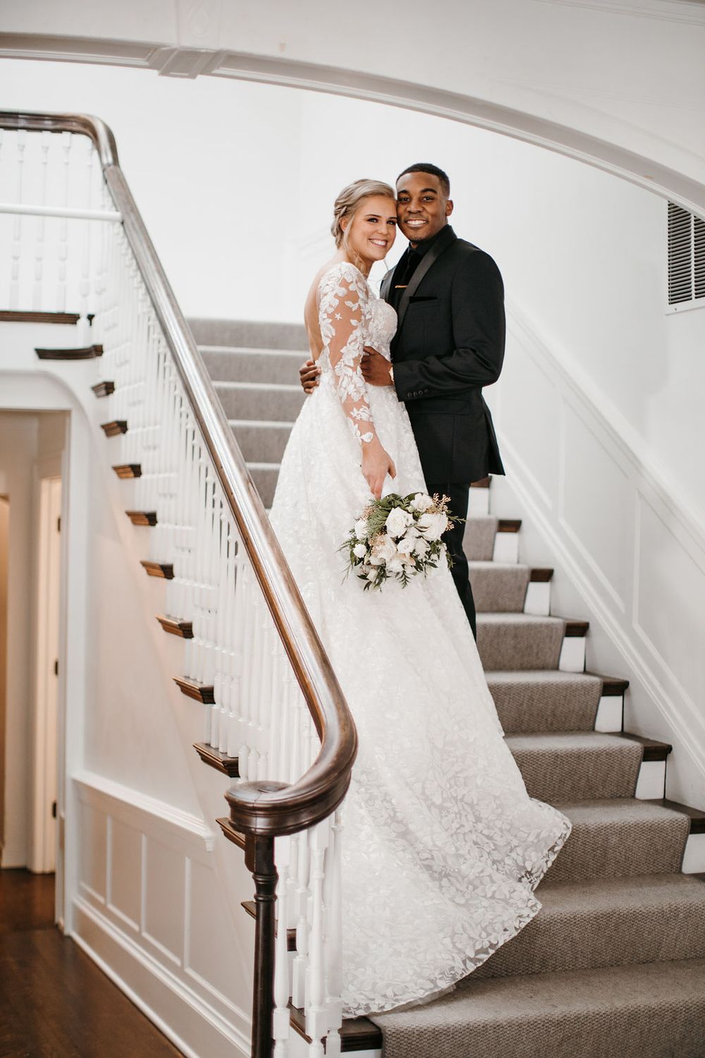 Bride and groom posing on elegant staircase, bride in lace gown holding white floral bouquet.